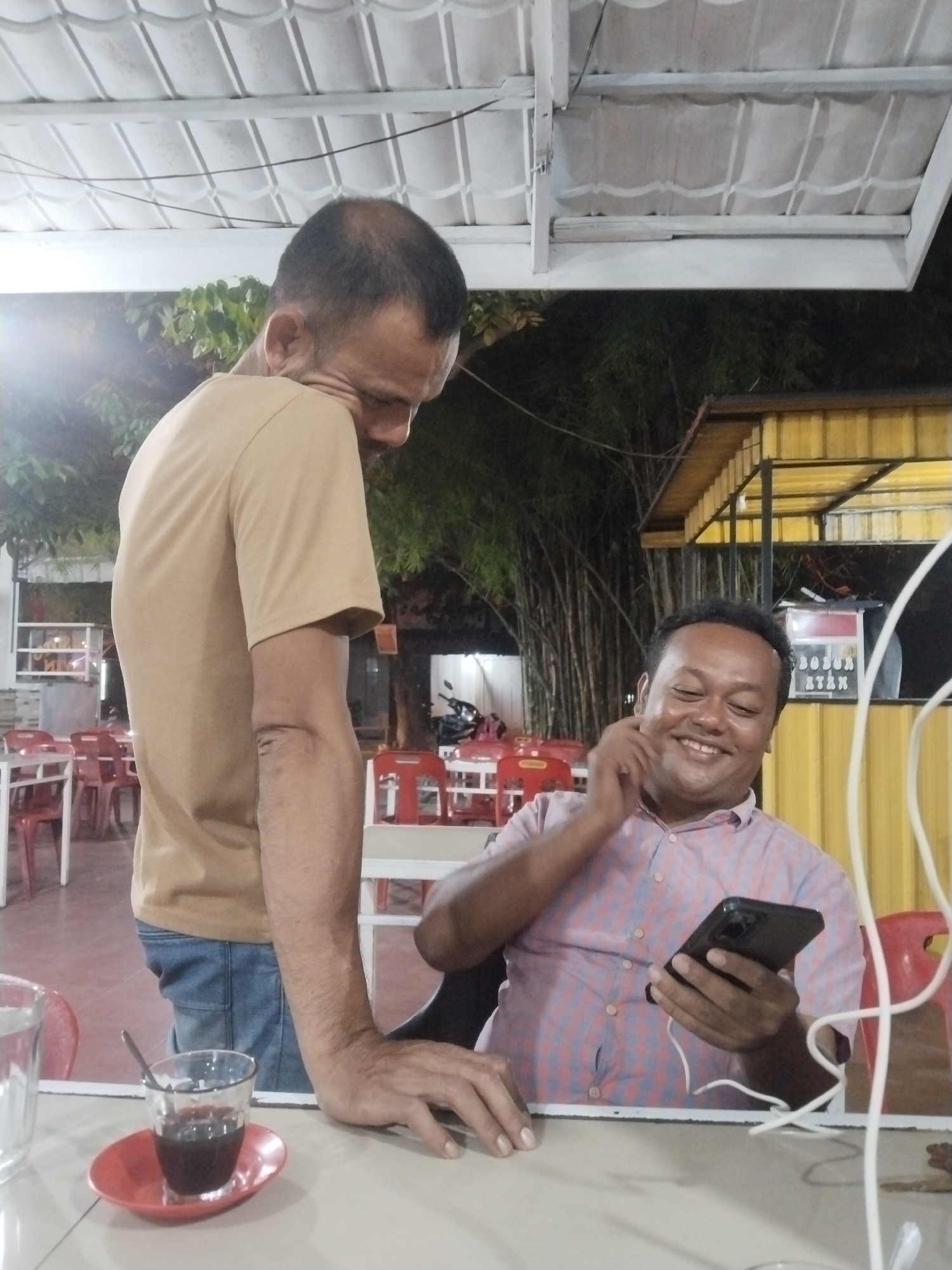 The image shows two men in an outdoor setting, presumably in Lhokseumawe, Indonesia, given the prompt. One man is standing, wearing a tan t-shirt and jeans, looking down towards the other man. The seated man is wearing a light pink and blue checkered shirt and is holding a smartphone, looking at the screen with a smile. He is also touching his ear. A small glass with a dark liquid and a spoon on a red saucer is on the table near the standing man. The surroundings appear to be a casual outdoor dining area with red plastic chairs and tables. In the background, there's a building with a yellow exterior and possibly bamboo trees. The lighting suggests it is either late afternoon or evening.