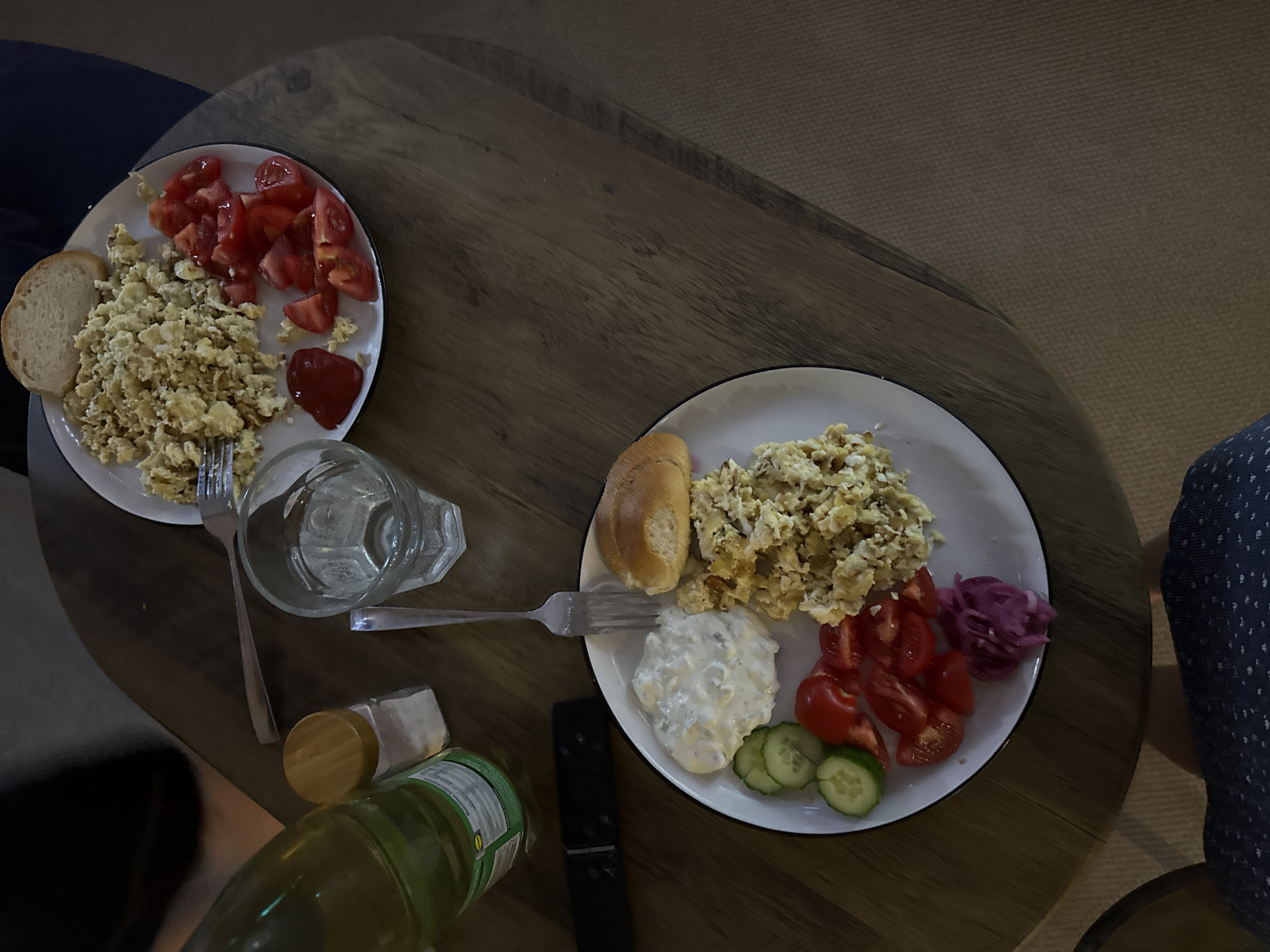 The image depicts a high-angle view of two plates of food placed on a dark brown wooden table. The left plate contains scrambled eggs, sliced tomatoes, a slice of bread, and a small amount of ketchup. The right plate holds scrambled eggs, a bread roll, a white creamy sauce, sliced tomatoes, sliced cucumbers, and pickled red cabbage. Each plate has a fork. There's a glass of water, a small container with a yellow lid, a bottle with a green label, and a black remote control. Parts of two individuals are visible on either side of the table; one wearing dark clothing and the other wearing a dark blue patterned garment. The scene is indoors. There is no information or indication that this is in Great Yarmouth, United Kingdom.