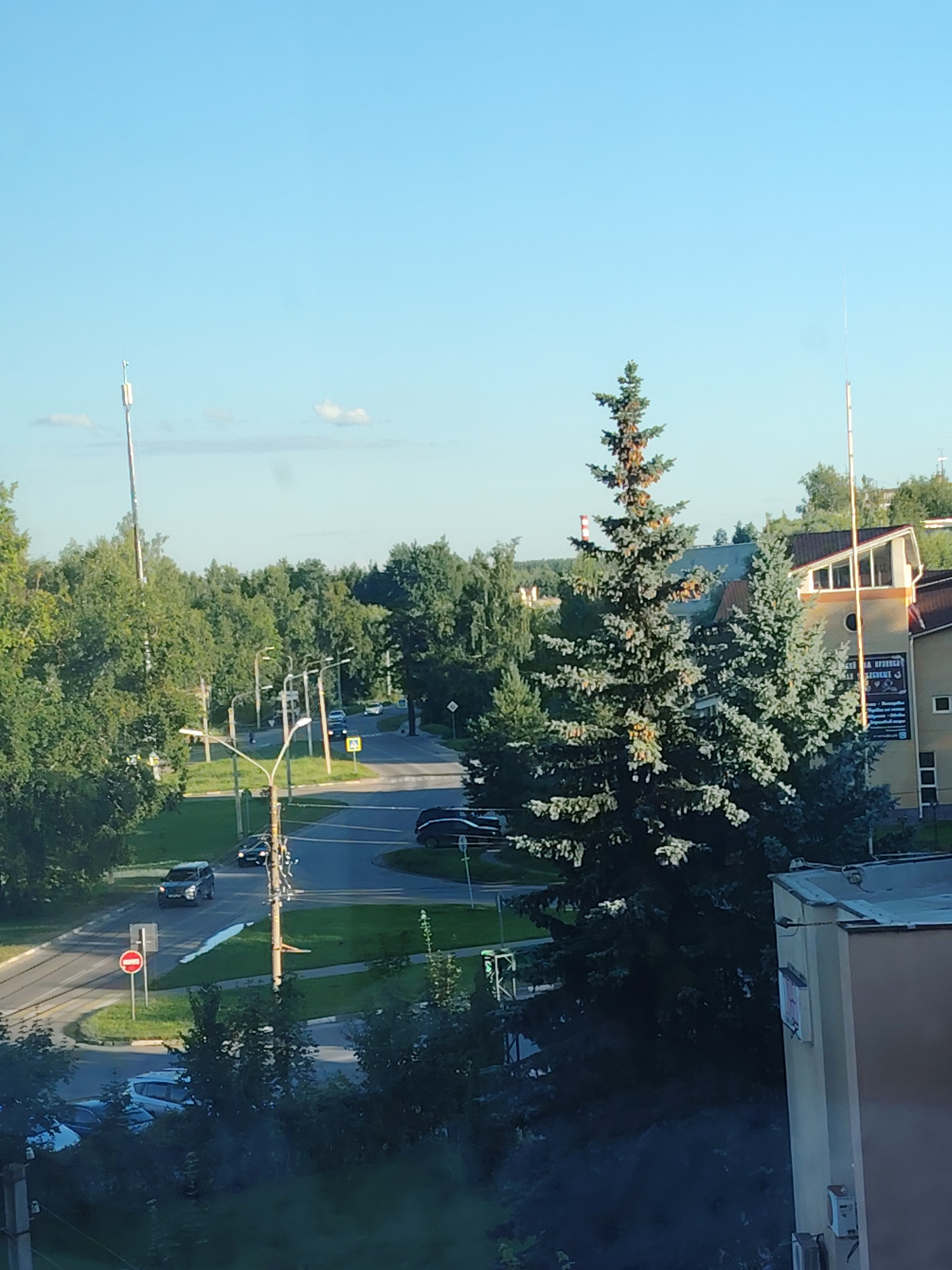 The image depicts an aerial view of a street scene in Konakovo, Russia, with a focus on a residential area interspersed with greenery and infrastructure. The foreground is dominated by the dark foliage of trees, obscuring the lower portion of the frame. A building, likely an apartment block, is visible on the right, its beige facade partially illuminated by sunlight. The building features a slanted roof, windows, and an air conditioning unit.
The middle ground reveals a road network with several vehicles, including a black car and a white SUV parked on the side of the road, and a black car in motion on the street. A red traffic sign with a white horizontal bar is positioned at an intersection. Streetlights are present along the roads, and the area is characterized by lush green grass and trees.
In the background, a dense forest stretches across the horizon, with the tops of trees visible under a clear blue sky. Two tall, slender towers, possibly communication or utility poles, are prominent features, one on the left and another on the right side of the image. The overall impression is one of a suburban or semi-urban environment during daylight hours, with a clear sky and pleasant weather.