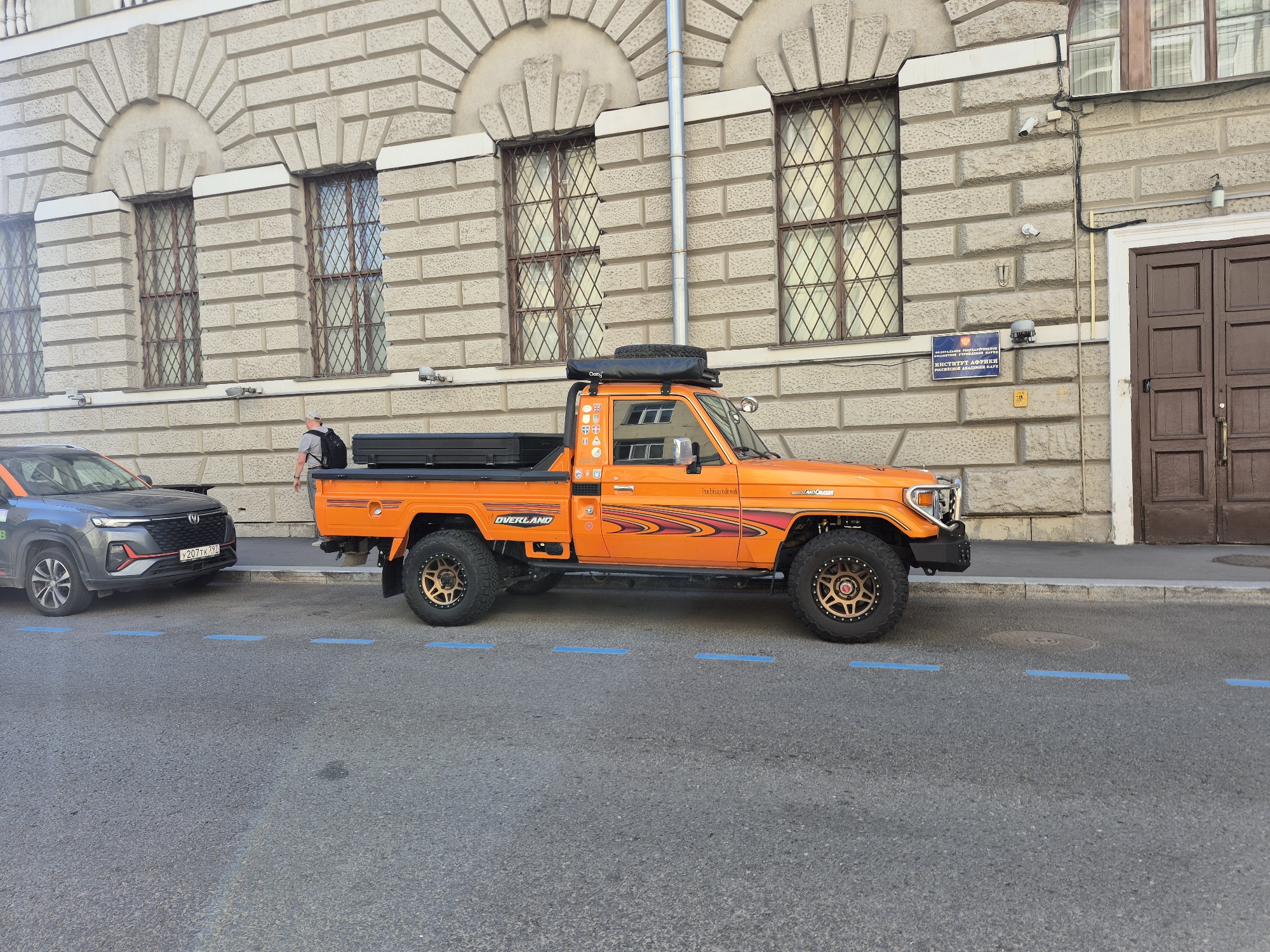 The image depicts an orange off-road vehicle, a Toyota Land Cruiser, parked on a street in Moscow, Russia. The vehicle is modified with an elevated roof rack, a spare tire, and aftermarket wheels. A black storage box is secured to the roof rack. A person wearing a grey t-shirt, grey cap, and a black backpack stands behind the vehicle. A dark grey SUV is parked to the left of the Land Cruiser. The building behind the vehicles has an ornate facade with multiple windows featuring diamond-shaped grilles. A sign on the building reads "ФЕДЕРАЛЬНОЕ ГОСУДАРСТВЕННОЕ БЮДЖЕТНОЕ УЧРЕЖДЕНИЕ НАУКИ ИНСТИТУТ АФРИКИ РОССИЙСКОЙ АКАДЕМИИ НАУК," indicating it is the Institute of Africa of the Russian Academy of Sciences. The street has blue parking markings.