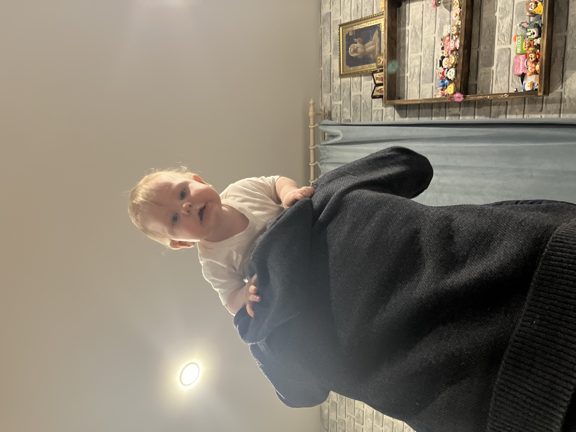 A baby is being held up by a person wearing a dark blue or black jacket. The room has a light-colored ceiling with a light fixture and a wall with a brick pattern. On the wall are two framed pieces of art and a wooden shelf with various objects on it. The location is Vesyoloye, Russia.