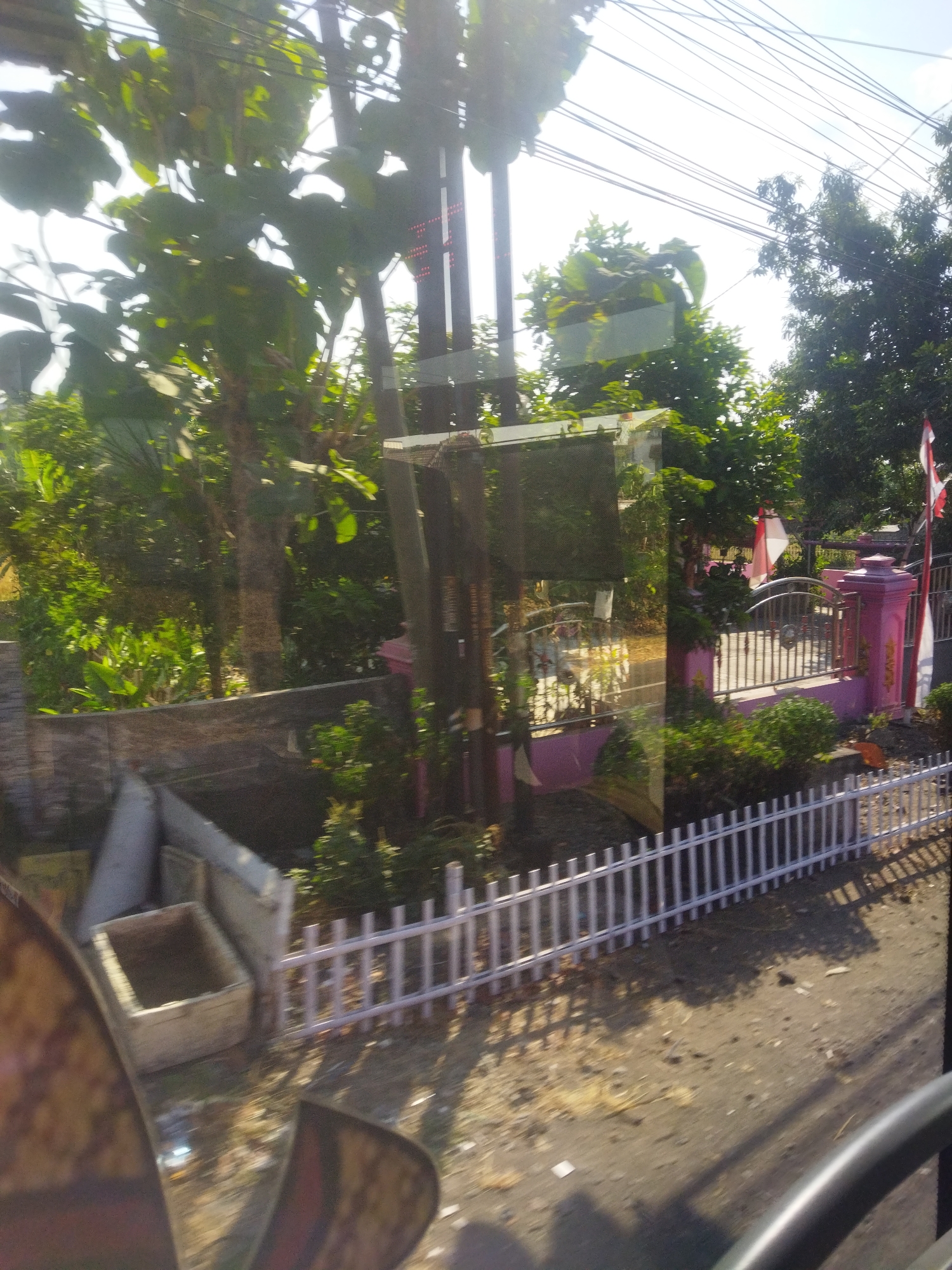 The image depicts a view from a moving vehicle in Selogiri, Indonesia. In the foreground, a portion of the vehicle's interior is visible, including a curved dark surface which appears to be part of the dashboard or steering wheel. The middle ground shows a white picket fence bordering a dirt path. Beyond the fence, there is a pink structure with decorative pillars and a metal gate. Lush green trees and vegetation are abundant throughout the scene. Two Indonesian flags are visible, one prominently displayed on a pole next to the pink structure and another smaller one further back. The sunlight is strong, creating shadows and highlights on the ground and foliage. The overall impression is of a residential or communal area in a tropical setting.