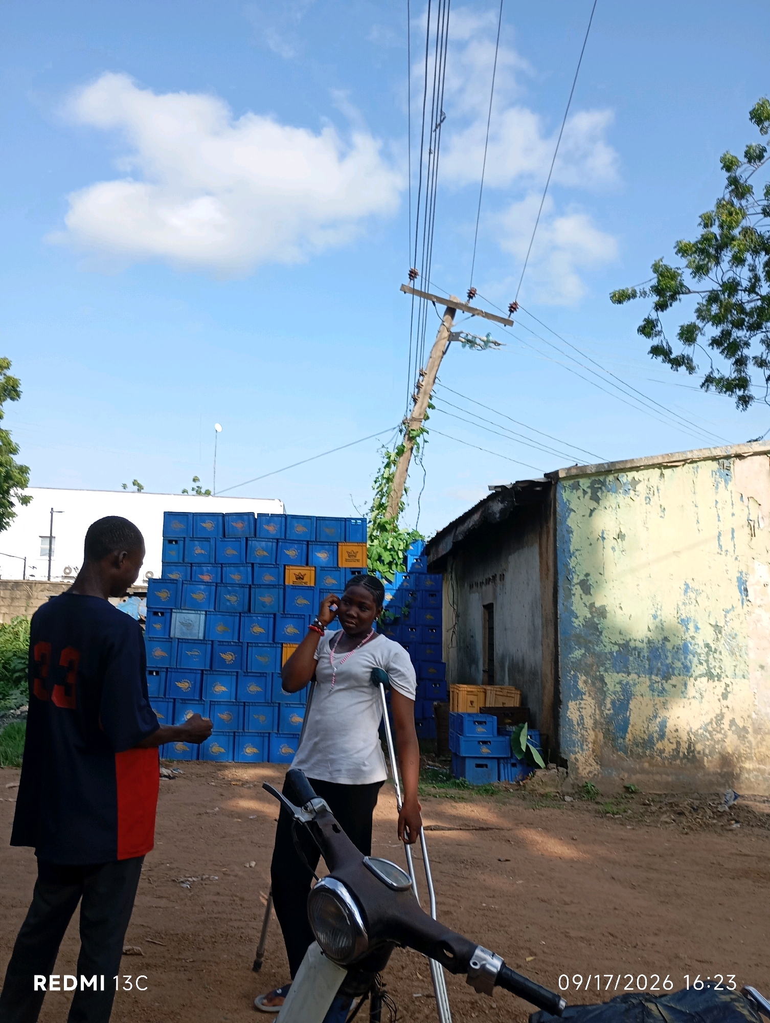 In Shendam, Nigeria, two individuals are present in an outdoor setting. A man, facing away from the camera, wears a dark blue t-shirt with a red stripe on the side and a red number "33" on the back. He stands to the left of the frame. To his right, a woman, who appears to be on a phone call, is using crutches. She is dressed in a white top and black pants. In the background, a large stack of blue crates, some of which are yellow, is visible, along with a dilapidated building with peeling paint. Overhead, power lines and a wooden utility pole dominate the sky, which is partly cloudy. A scooter is partially visible in the foreground. The ground is covered in dirt and gravel.