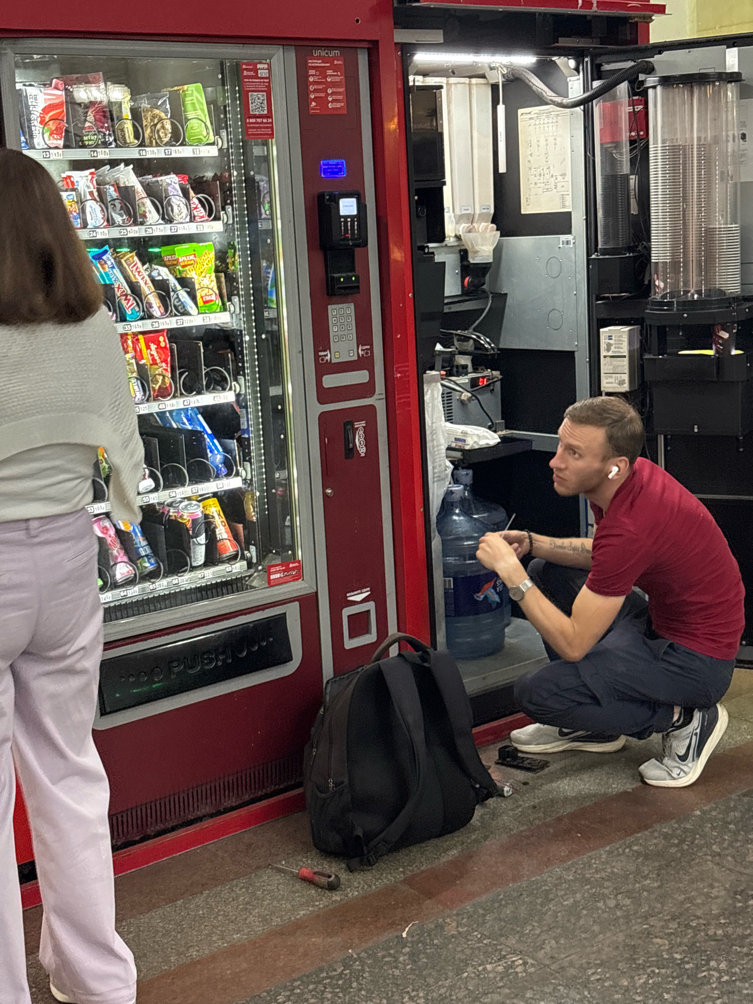A man in a red shirt and dark pants squats next to a vending machine and two large water jugs. He is looking to the right, wearing white earbuds and a watch. In front of him, a black backpack rests on the floor. A small screwdriver lies on the ground to the left of the backpack. To the left of the man and jugs, a woman in a white sweater and light purple pants stands facing away from the camera, looking at the vending machine. The vending machine is red and displays various snacks and drinks. The floor is a textured speckled gray and red material. The scene appears to be in an indoor public space, possibly a subway station or a transit hub in Moscow, Russia, given the "Unicum" branding on the vending machine which is a Russian brand.