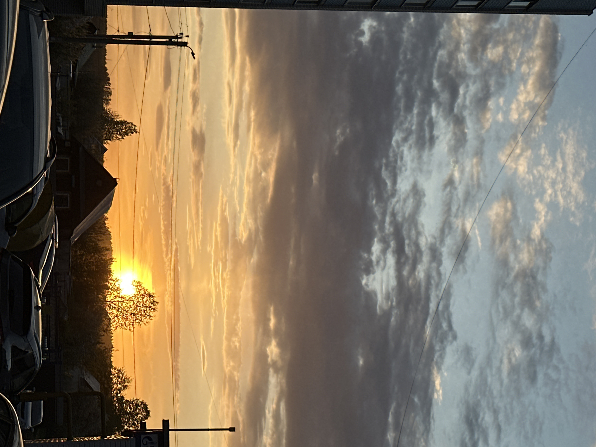 The image captures a sunset view in what appears to be an urban area in Chaikovsky, Russia. The sun is setting behind trees and a building, casting a golden glow on the sky and clouds. The clouds are a mix of golden hues and grays, creating a dramatic effect. Utility poles and wires are visible against the sky. In the foreground, parked cars are partially visible. The top of a building, possibly an apartment complex, is seen at the top of the image. The image composition is slightly rotated clockwise.