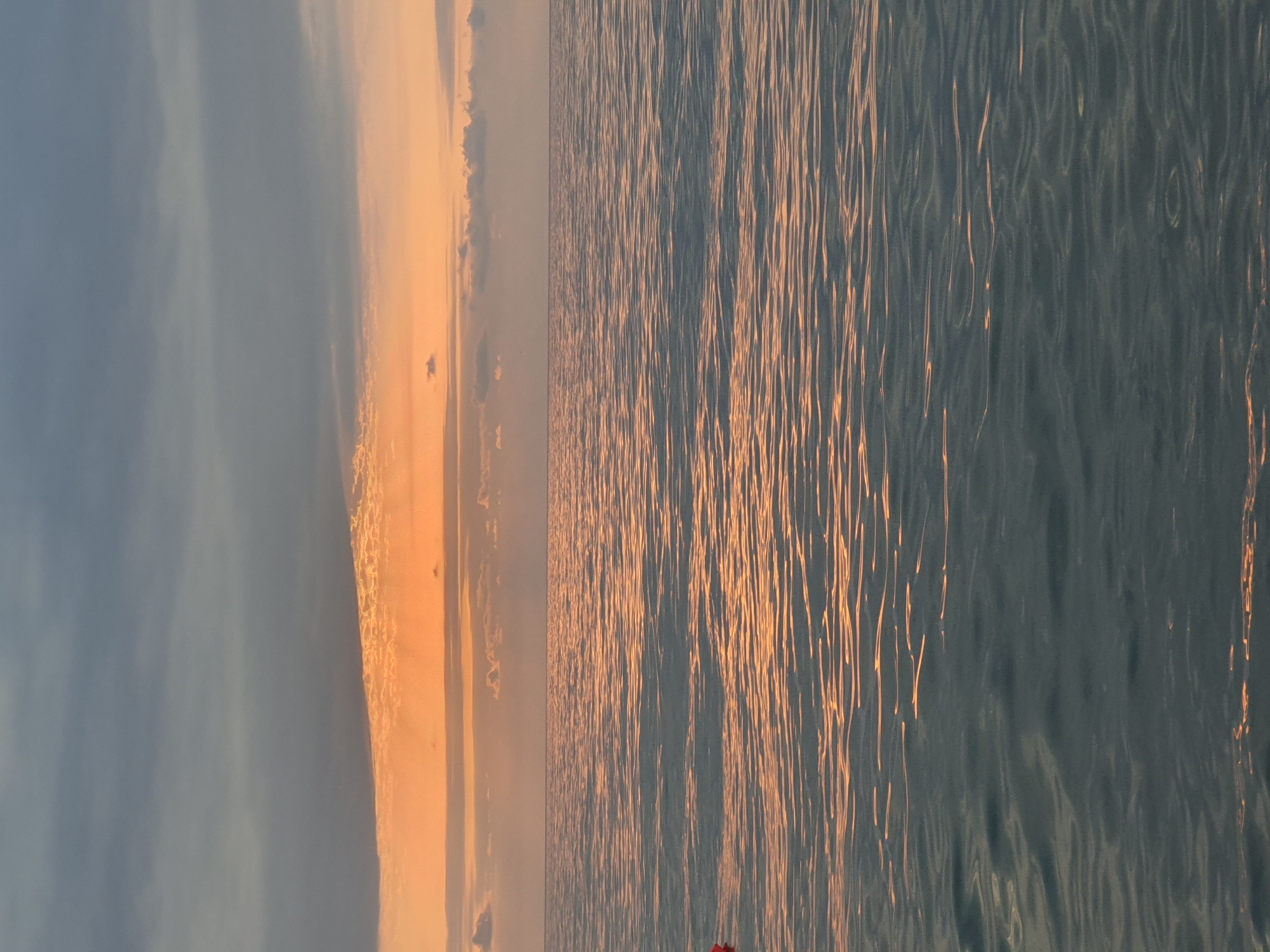 A vertical, close-up shot captures the vast expanse of the sea under a dramatic sunset sky. The water in the foreground is a deep, textured blue-grey, with subtle ripples catching the fading light and reflecting streaks of orange and pink. The horizon line is sharp, dividing the sea from the sky. The sky is a gradient of colors, transitioning from a soft orange glow near the horizon, where thin, wispy clouds are illuminated by the setting sun, to a darker, dusky blue-grey overhead. A small, red object, possibly a buoy, is partially visible on the far left edge of the frame, resting on the water. The image evokes a sense of calm and the quiet beauty of nature. The location is the Black Sea coast in Batumi, Georgia.