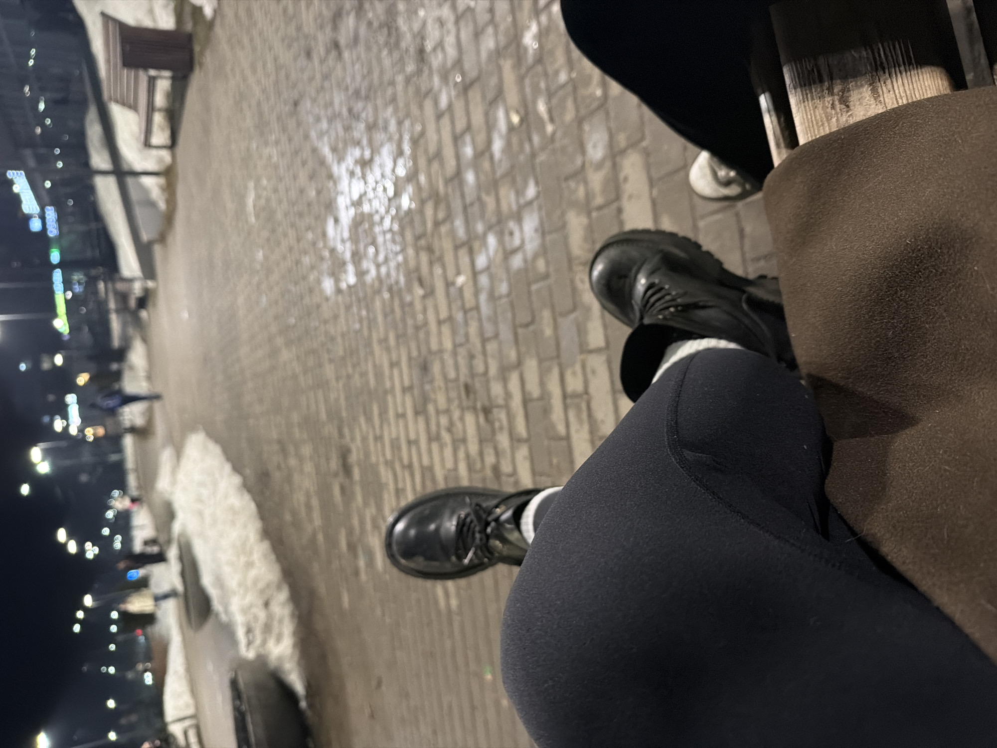 The image shows a first-person perspective of someone sitting on a bench in Chaikovsky, Russia. The person is wearing black leggings and black boots with white socks. Another person can be seen on the bench next to the first person, wearing black pants and white shoes. The bench has wooden slats and a brown armrest. The ground is made of interlocking pavers with patches of snow. In the background, there are streetlights and buildings. The photo was taken at night.