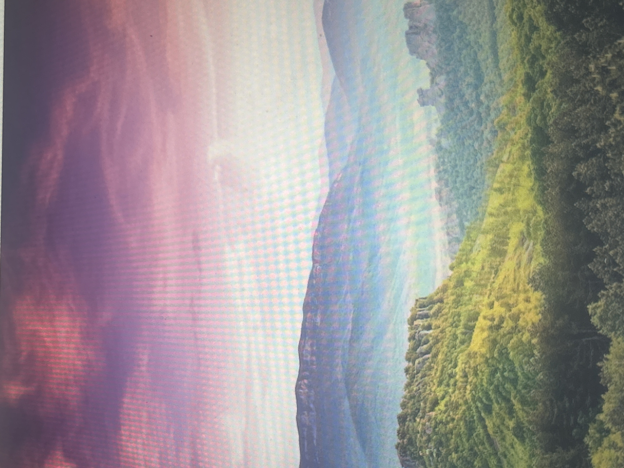The image depicts a majestic mountainous landscape, bathed in the soft hues of a sunset or sunrise. The sky transitions from a deep, rich magenta at the top to a pale pink and then a light, ethereal blue as it meets the mountains. Wispy clouds stretch across the sky, catching the light and adding depth to the scene.
The foreground is dominated by a lush, green mountainside covered in dense forestation. Sunlight illuminates patches of the foliage, creating a striking contrast between light and shadow. A prominent, flat-topped rocky outcrop juts out from the mountainside, its rugged texture clearly visible. Further back, layers of misty mountains recede into the distance, their forms softened by the atmospheric haze and the fading light. The distant peaks appear as hazy blue silhouettes against the sky.
This specific mountainous landscape is identifiable as Vitosha Mountain, located on the outskirts of Sofia, Bulgaria. The distinctive rock formations and layered mountain ranges are characteristic features of Vitosha.