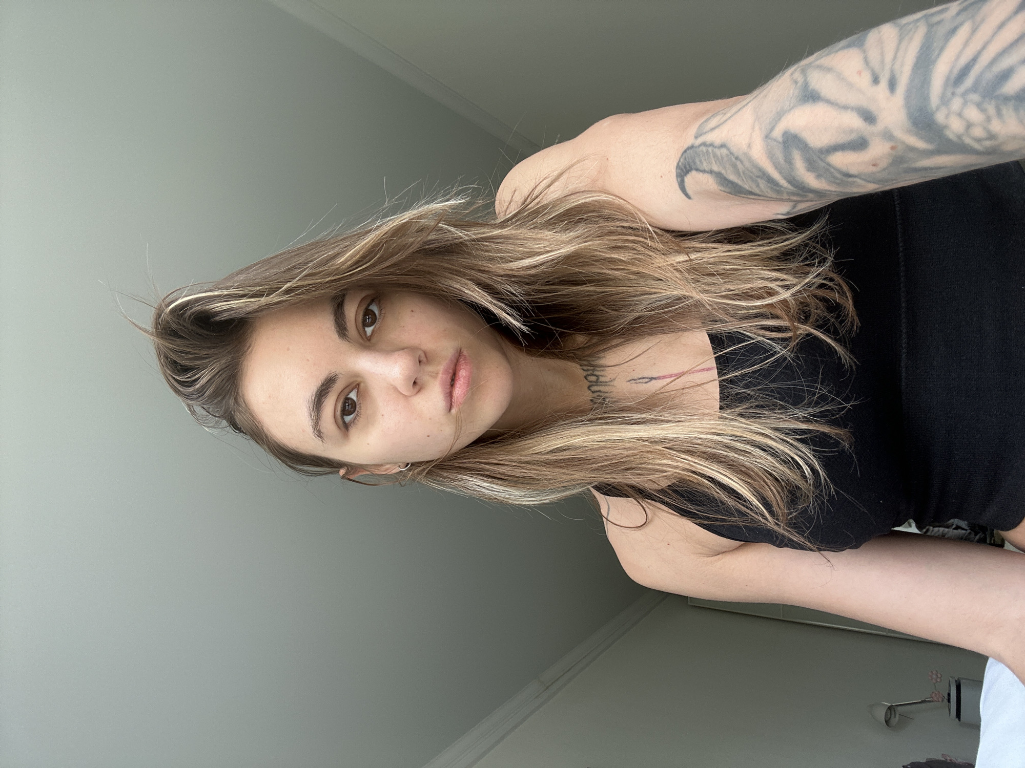 A young woman with long, wavy blonde and brown hair, wearing a black tank top, is looking upwards towards the camera. She has brown eyes and fair skin, with freckles visible on her face. A tattoo is visible on her left arm, and another tattoo is partially visible on her chest. The background is a plain, light grey wall and ceiling. The image appears to be taken indoors.