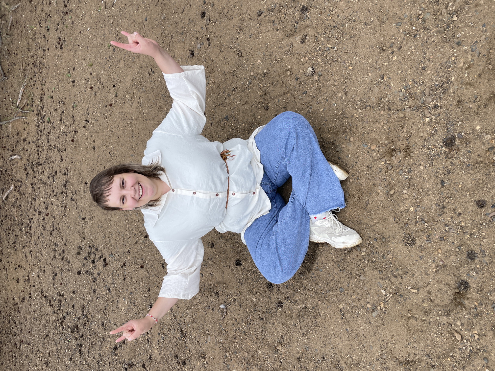 A woman is sitting cross-legged on the ground. She is wearing a white, oversized button-up shirt, blue wide-leg jeans, and white sneakers with red accents. A thin brown belt with a tassel detail cinches her waist. She has short, brown hair and is smiling at the camera, holding up two fingers in a peace sign with each hand. The ground appears to be dirt with scattered pinecones and small stones. The location is the city of Chaikovsky, Russia.