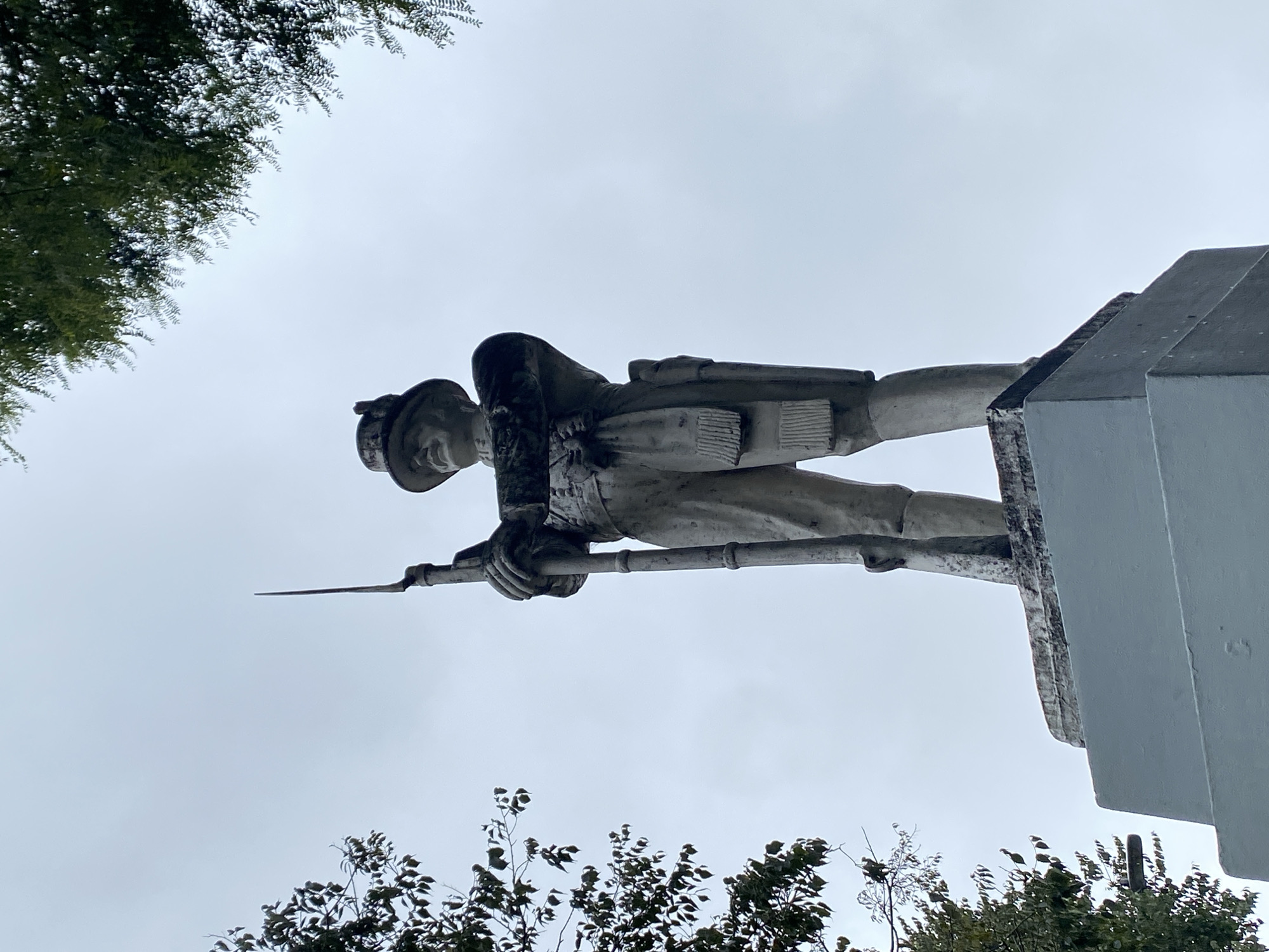 The image displays a weathered statue of a soldier, depicted in a monochrome tone suggesting stone or concrete, viewed from a low upward angle against an overcast sky. The statue, standing upright, portrays a soldier in a military uniform and helmet or cap, holding a rifle with a fixed bayonet. His hands grip the rifle, the butt of which would rest on a surface below. Signs of age and environmental exposure, such as dark stains or discoloration, are visible on the statue, particularly on the soldier's back and parts of the robust, block-like pedestal to the right. The sky is a uniform light grey, characteristic of a cloudy day with soft, diffused lighting. Green foliage from trees partially frames the scene in the upper left and lower right corners. As a static monument, no active event or activity is taking place. There is no visible text on the statue or in the background. The monument is situated in Buenos Aires, Argentina.