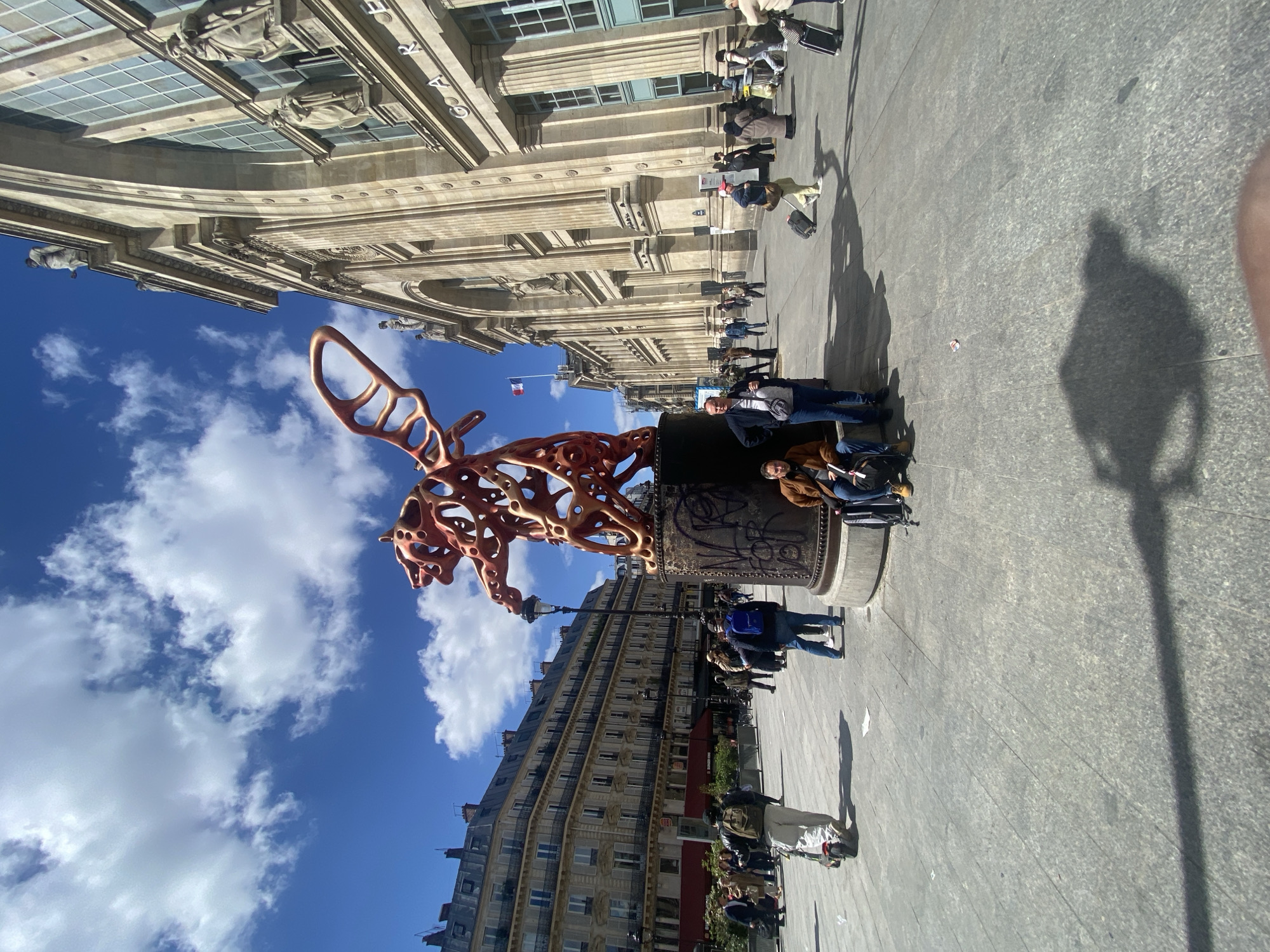 The media file captures a vibrant outdoor scene in Paris, France, under bright daylight. In the foreground and midground, a striking reddish-orange, abstract sculpture of an animal, possibly a rooster or a lion, crafted with an open, organic, and intricate design, stands atop a dark, cylindrical pedestal. The pedestal itself is adorned with etched patterns and some visible graffiti.
Several people are present in the wide, paved public square. Two individuals are seated on the base of the sculpture, while others walk or stand nearby. Many are seen with luggage, suggesting they might be travelers or tourists. Strong, elongated shadows cast across the light grey flagstone pavement indicate a sunny day, likely midday or early afternoon.
In the background to the right, a grand, multi-story building with classical architectural elements, such as columns, cornices, and statuary, dominates the skyline. The word "GAR" is partially visible on its facade, hinting at a "Gare" (train station). A French flag flies from a pole near this building. To the left, another multi-story building with characteristic Parisian architecture, featuring rows of windows and balconies, lines the street.
The sky is a clear blue with scattered, fluffy white clouds, contributing to the bright and inviting atmosphere. The overall scene depicts a bustling urban environment, with people moving about amidst impressive architecture and modern art.