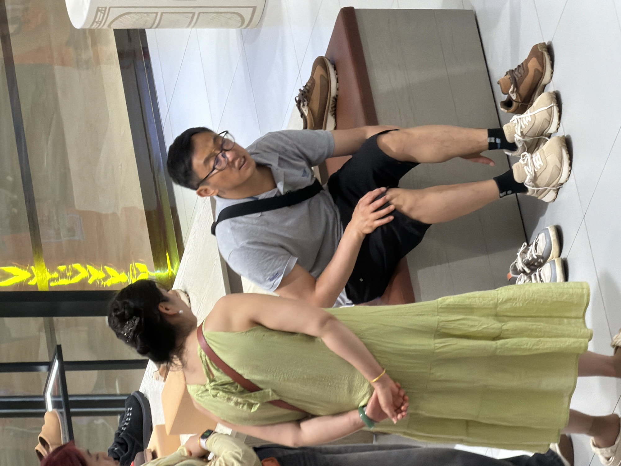 In a well-lit shoe store, possibly located in Yuzhong District, China, a man wearing glasses, a grey polo shirt, and black shorts sits on a padded bench, looking down. To his right, a pair of brown and tan athletic shoes rest on the bench, and another similar pair is on the floor in front of him. A woman in a lime-green, tiered dress stands to the left, her back to the camera, hands clasped behind her. A maroon strap from her bag is visible across her back. Several other pairs of shoes are visible on display shelves in the background.