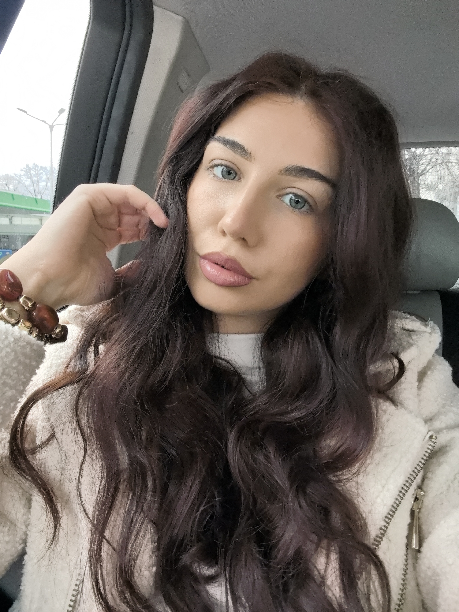 The image displays a female subject seated inside a vehicle in the city of Stara Zagora, Bulgaria. The subject, positioned centrally and looking directly at the camera, has long, wavy dark brown hair and light blue eyes. She wears a white top under a cream-colored sherpa jacket with a visible zipper. A multi-beaded bracelet, featuring large reddish-brown oval beads interspersed with smaller metallic gold-colored beads, is present on her left wrist. Her left hand is raised, with fingers lightly touching her hair near her left temple. The vehicle's interior visible elements include a dark gray seat headrest behind her and a light gray ceiling. Through the front windshield, an overcast sky is visible, along with a multi-armed street light pole on the upper left. A horizontal green structure is partially visible in the mid-ground, and bare deciduous trees are discernible in the distant background. The scene is illuminated by diffuse natural light.