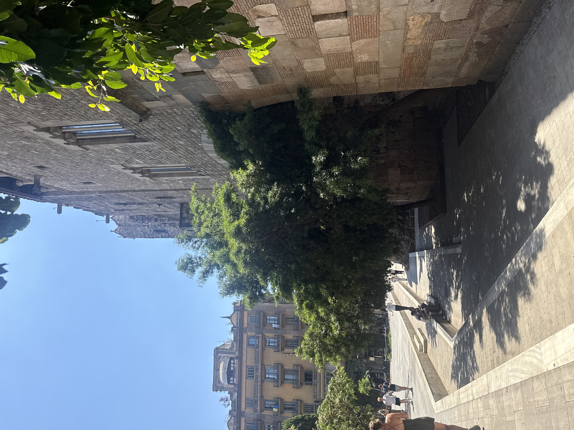 The image depicts an outdoor scene in Barcelona, Spain, on a clear, sunny day. The perspective is unusual, with the bottom of the image appearing at the top.
In the foreground, lush green foliage partially obscures a view of a building and an outdoor staircase. The building appears to be old, with a facade made of brick and stone. Several windows are visible, some with shutters.
A wide, paved staircase with distinct light-colored steps dominates the right side of the image, leading upwards and to the left. Shadows from trees are cast across the steps, indicating a time of day when the sun is high in the sky, likely midday.
Several people are scattered on the staircase and surrounding area. Some are walking, while others are seated or gathered in small groups, suggesting a casual, leisurely atmosphere. A few individuals are dressed in casual summer attire.
Further in the background, more buildings are visible, characteristic of Spanish architecture with their balconies and window arrangements. The sky is a clear, vibrant blue. The overall impression is one of a bright, warm day in an urban environment with significant historical architecture and vibrant plant life. There is no visible text in the image.