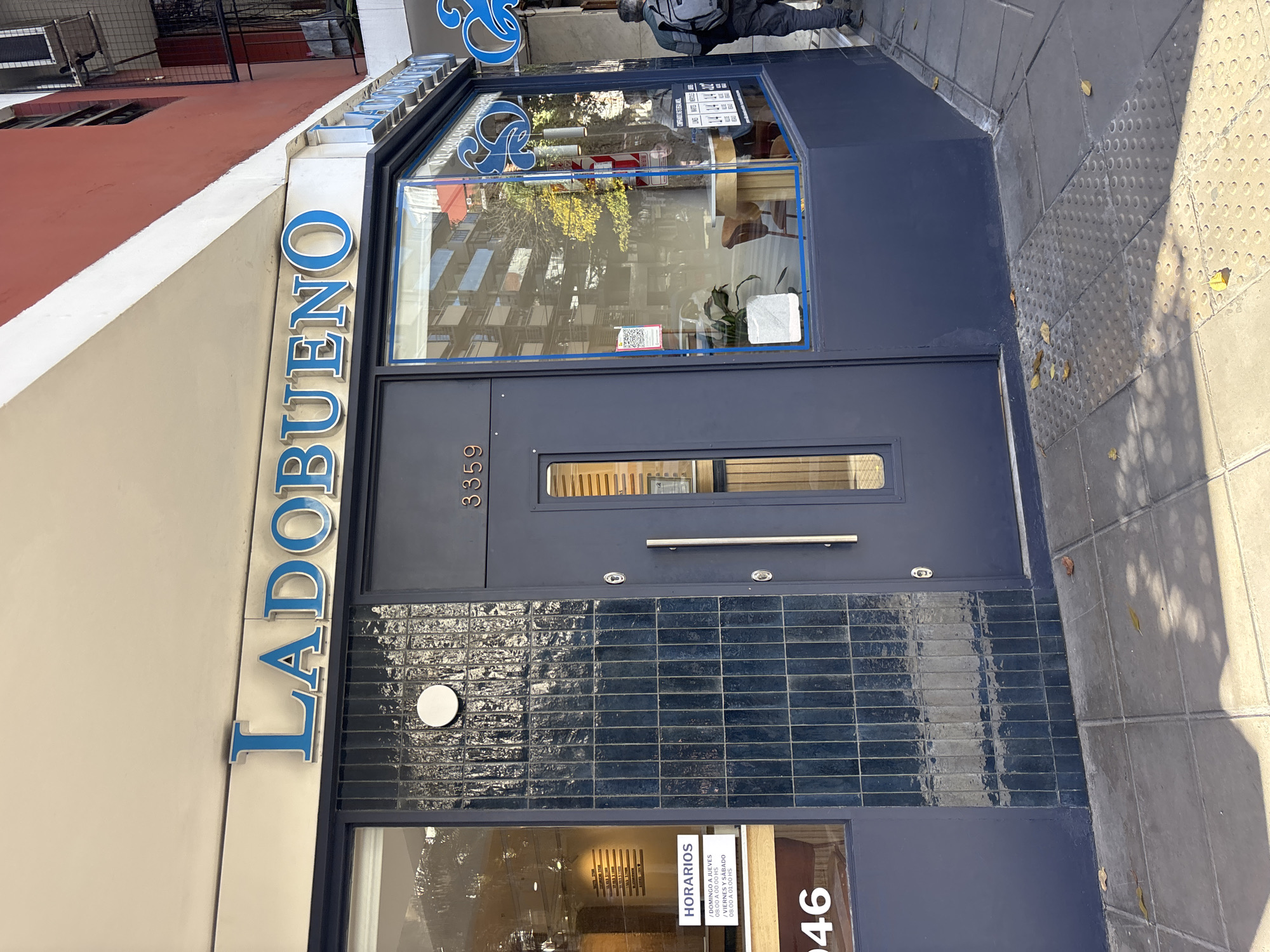 The image shows the facade of a building in Buenos Aires, Argentina. The sign vertically spells out "LADOBUENO" in blue lettering on the light-colored wall of the building. The front door is a dark blue, with the number "3359" inscribed above the door. Below the door handle and peephole, there are glazed, dark blue, rectangular tiles, arranged horizontally. A smaller glass window below reveals an opening hours notice written in Spanish, and partially shows the number "46". The sidewalk is gray, with a few fallen leaves. A person in dark clothing can be partially seen on the upper right.