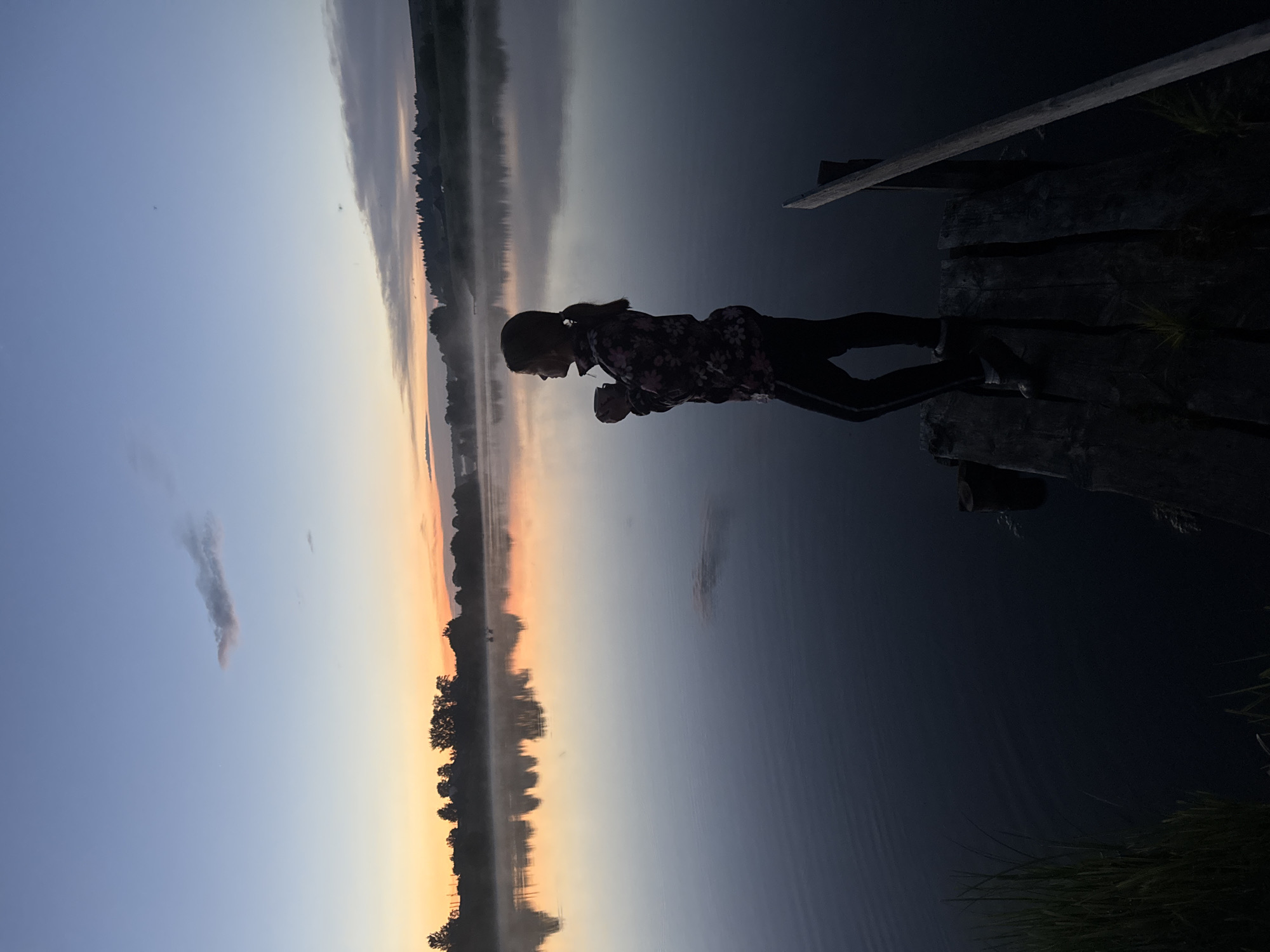 The image depicts a serene dawn scene at a lake, presumably in Amaneevo, Russia, as suggested by the provided context. A young person, silhouetted against the softly lit sky, stands on a wooden pier. They are holding a cup, appearing to enjoy the quiet morning. The water of the lake is calm, reflecting the colors of the sunrise – soft oranges, pinks, and blues – and the silhouettes of trees lining the distant shore. A gentle mist hovers over the water's surface, adding to the tranquil atmosphere. The sky transitions from a pale blue at the top to warmer hues near the horizon, with a few wispy clouds present. The pier itself is made of weathered wooden planks. The overall impression is one of peaceful solitude amidst the natural beauty of a lakeside dawn.