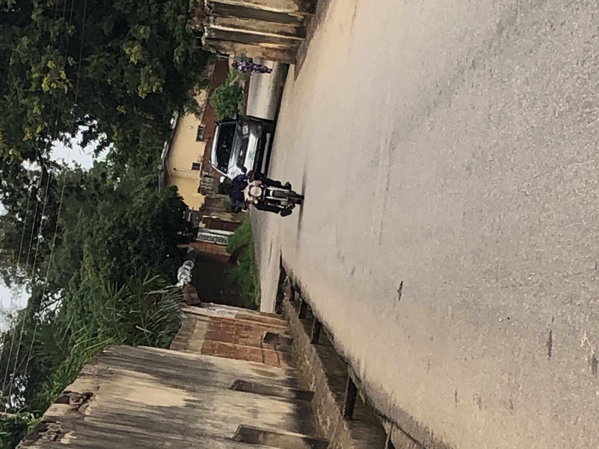 A man rides a motorcycle down a paved road, with a silver SUV behind him and a woman in a colorful dress walking away from the motorcycle and SUV on the right side of the road. The road is flanked by walls on both sides, with vegetation growing behind the walls and on the left side of the road. The scene appears to be in a residential area, with a house visible in the background. The weather appears to be overcast.