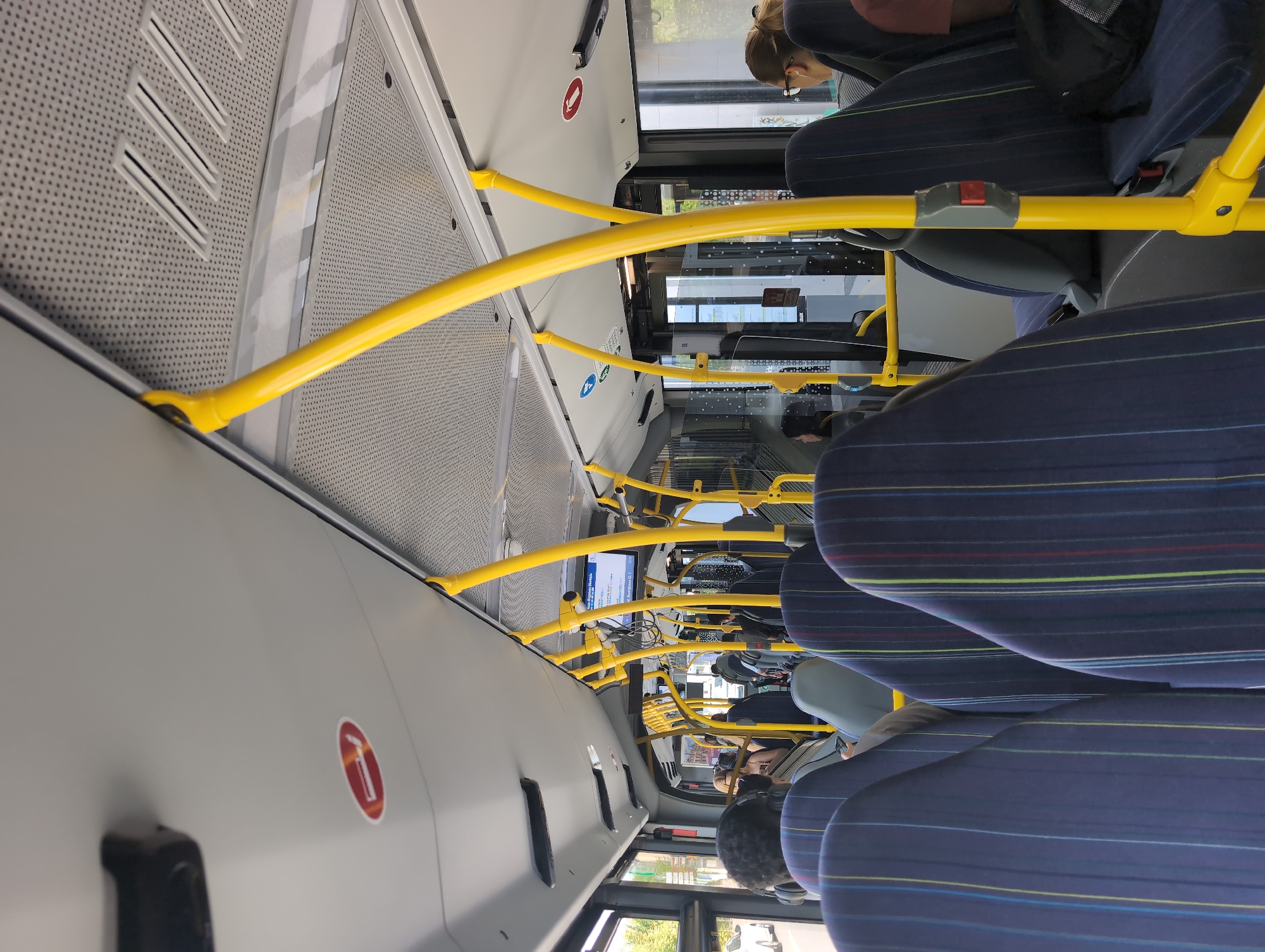The image shows the interior of a bus. The seats are upholstered in dark blue fabric with thin vertical stripes of yellow, green, and red. Yellow metal poles are visible throughout the bus, serving as handrails. The ceiling is white with a patterned texture and contains air vents and lighting fixtures. Several passengers are seated on the bus, mostly seen from the back or side. Some passengers are wearing headphones. A digital display board is mounted above the seats towards the front of the bus. There are also various warning and informational signs on the ceiling, including a red circular sign with a diagonal line through a cigarette, indicating no smoking. The overall environment suggests a public transportation setting on a bright day, as evidenced by the natural light coming in from the windows.