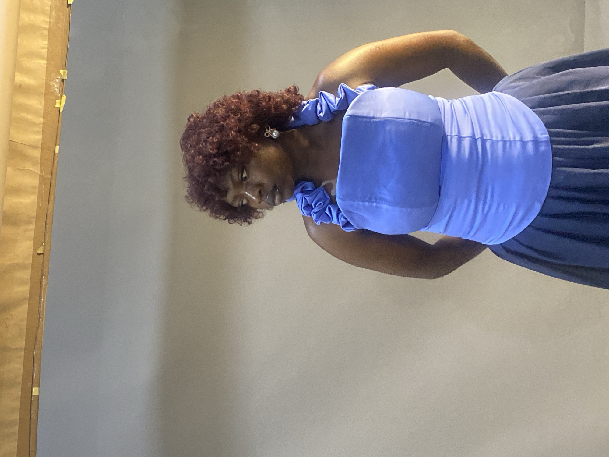 A woman is posing for a picture in what appears to be a studio setting. She is wearing a blue dress with ruffled shoulder straps and a voluminous skirt. Her hair is curly and a reddish-brown color. She has a pearl earring in her left ear. Her head is tilted down, and she appears to be looking at something below her. The background is a plain, light-colored wall. To the left of the woman, there is a large object covered in brown paper that is taped to the wall. The lighting suggests it is daytime, but no other weather or location cues are present. The emotion on the woman's face is neutral. There is no visible text in the image.
