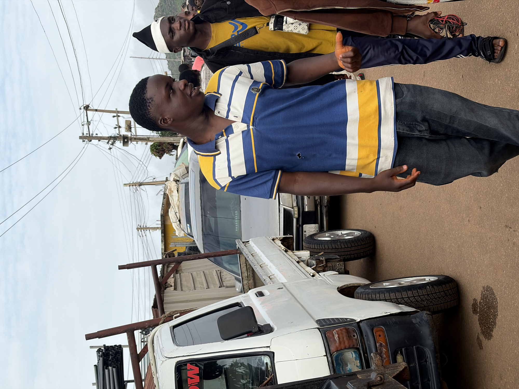 In Gura Top, Nigeria, on a cloudy day, a young man in a blue and yellow striped polo shirt and dark jeans stands holding a small, orange object, possibly a carrot, in his right hand. He is facing towards the left of the frame and appears to be speaking. To his right, another young man wears a yellow t-shirt and a black jacket, with a white and black hat. Behind them, a white flatbed truck is parked. In the background, power lines crisscross the sky, and a few buildings are visible. The ground is a dirt road with some spilled liquid. The scene suggests a market or roadside setting.
