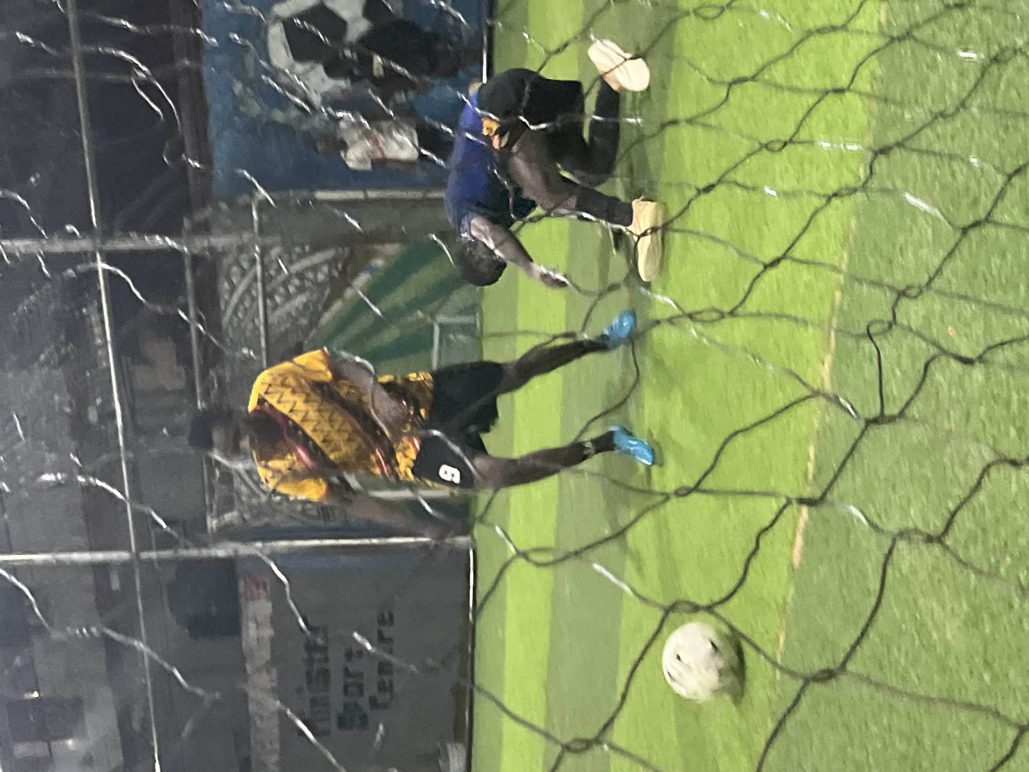 The image depicts a low-light, indoor futsal court. Two young men are visible on the green artificial turf. One man, wearing a yellow and black patterned jersey with the number 9 on his shorts, is in motion, his legs bent as if running or kicking. He is wearing bright blue athletic shoes. Another man, in a dark blue shirt and black pants, is on his knees on the ground, reaching forward with his right hand. He is wearing tan-colored shoes. A white soccer ball is visible on the turf to the left of the kneeling man. In the background, other individuals can be seen, some standing, and what appears to be a large soccer ball graphic on a blue wall. A sign with text, possibly "Minista Sport Centre," is visible on a grey structure. The scene is partially obscured by a black net in the foreground. The overall impression is of an active futsal game.