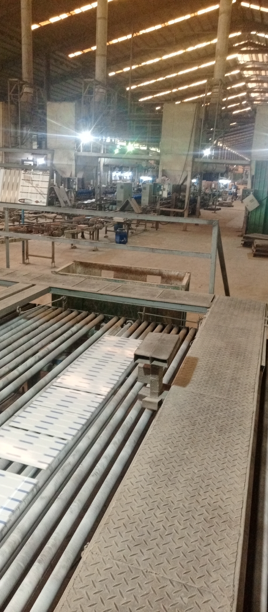 The image depicts an industrial setting, specifically a factory in Magboro, Nigeria. The scene is dominated by manufacturing equipment, including a roller conveyor transporting rectangular objects, likely tiles or panels. A metal walkway with a diamond-plate pattern runs alongside the conveyor. In the background, various machines and structures are visible, partially obscured by distance and haze. The overall lighting suggests a large indoor space with sunlight filtering through the roof structure. There are no people visible in the immediate foreground.