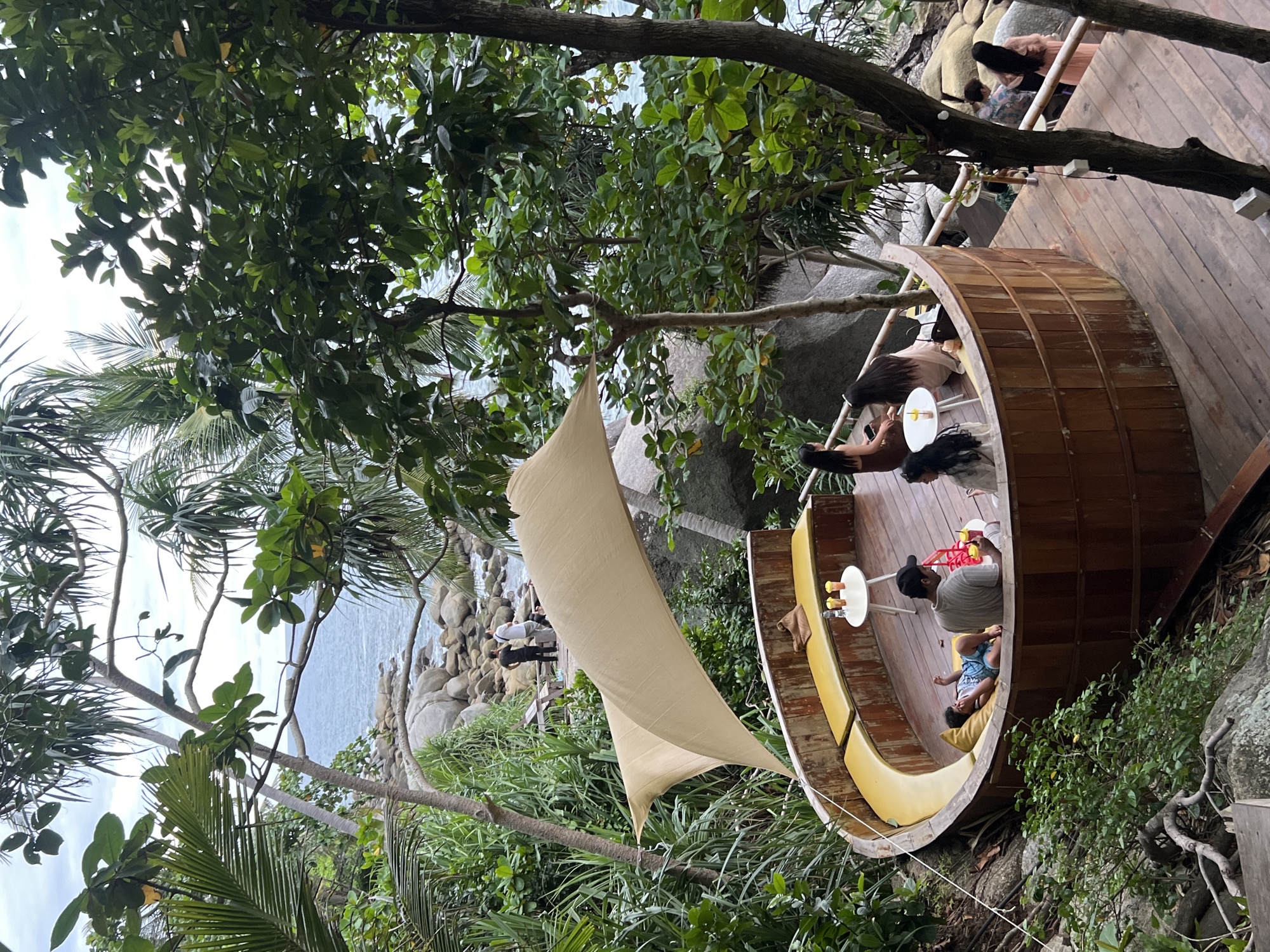A family is seen relaxing in a semi-circular wooden outdoor seating area at a restaurant with an ocean view in Karon, Thailand. The seating area has yellow cushions and a large, beige canopy providing shade. A young boy lies on a yellow cushion, appearing relaxed. A man and two women are seated on the cushions. There are small white tables with drinks on them. In the background, the ocean and a rocky shoreline are visible, surrounded by lush green vegetation and palm trees. Further in the distance, a few other people are seen standing near the shore. A wooden deck extends from the seating area, with a few more people visible on it. The overall scene depicts a tranquil and scenic dining experience.