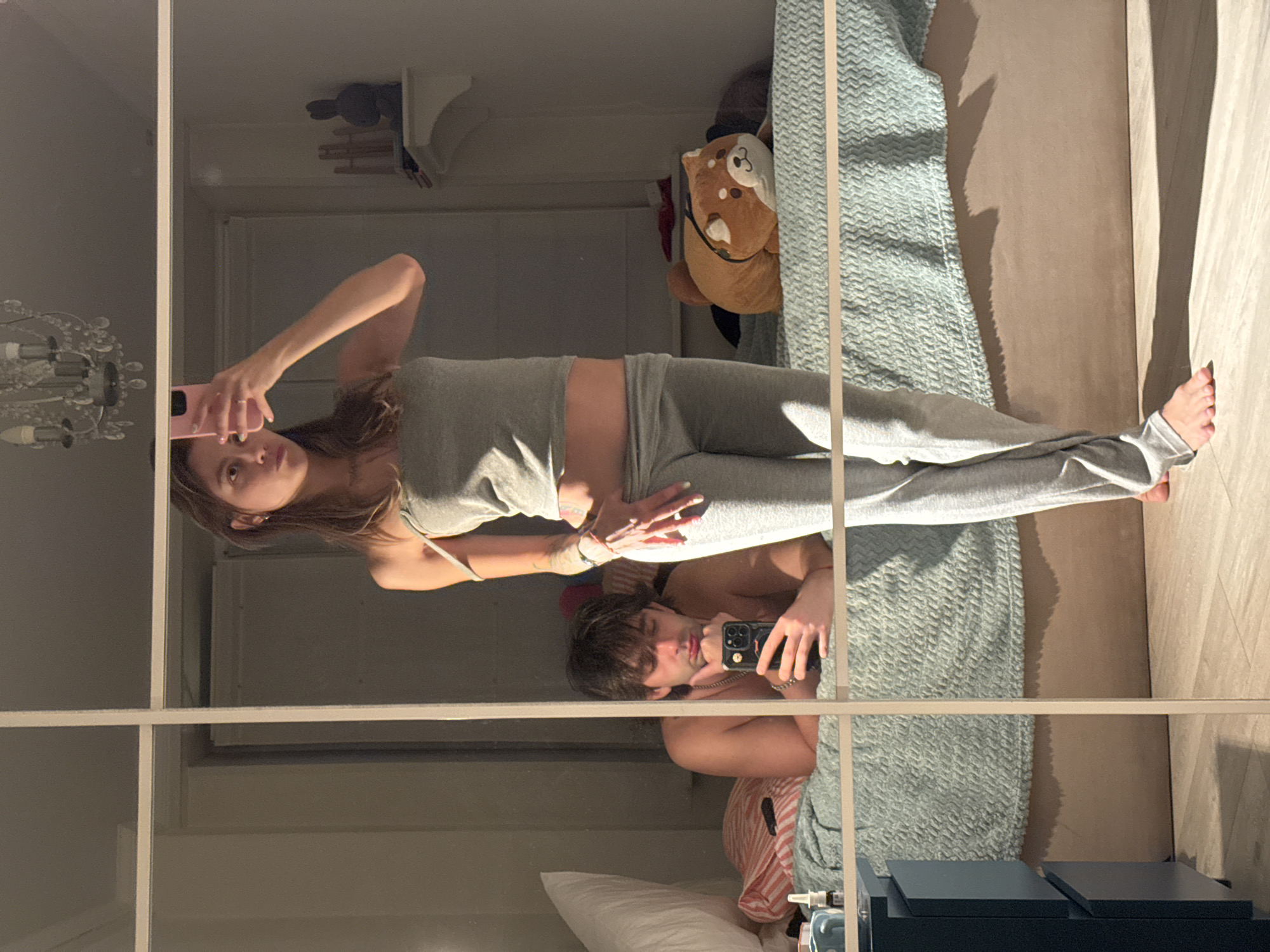The image shows a bedroom, likely in Moscow, Russia, as indicated by the user prompt. A young woman is taking a mirror selfie, holding a pink phone in her right hand. She is wearing a grey tank top and grey sweatpants. Her left shoulder strap is pulled down. She has long, brown hair and appears to be wearing makeup. She has a ring on her left hand. To her right, reflected in the mirror, is a shelf with decorative items, including a dark silhouette figure. A chandelier hangs from the ceiling, also reflected.
In the background, lying on a bed, is a young man. He is shirtless and wearing a silver chain. He is looking at his phone, which he holds in his left hand. He is resting his chin on his right hand. The bed has a light teal quilted blanket and striped pillows. A stuffed animal, resembling a Shiba Inu, is placed on the bed to the woman's right. A dark blue nightstand is visible to the left of the bed. The room is lit by natural light coming from the right, casting long shadows on the floor. The overall impression is that of a candid moment captured in a private residence.