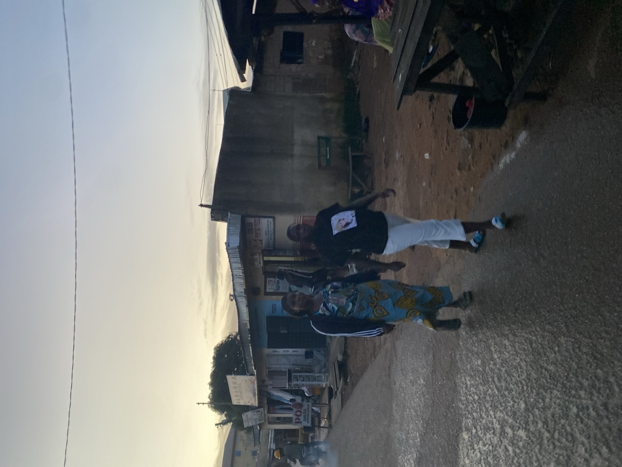 The image captures a street scene in Jos, Nigeria, during what appears to be dawn or dusk, indicated by the soft, diffused light and pale sky. In the foreground, a paved road with a wet, textured surface leads into the frame. Two women are walking side-by-side on the road. The woman on the left wears a long, patterned blue and yellow dress with a black jacket, and dark sandals. The woman on the right wears a black t-shirt with a portrait on it and white trousers, along with blue and white sandals.
Along the right side of the road, a rough, unpaved area is visible, with scattered debris and a dark bucket filled with red objects. A rustic wooden table or stall is set up, casting a shadow. The background features a row of low-rise buildings, likely shops, with corrugated iron roofs. Signs are visible, including one that reads "ALBARKA" and another with "POS DATA AVAILABLE". Further down the street, a person on a motorcycle, wearing a yellow hard hat, is visible, creating a slight dust trail. The overall impression is of a casual, everyday street in a Nigerian city, with ongoing commercial activity and pedestrian traffic.