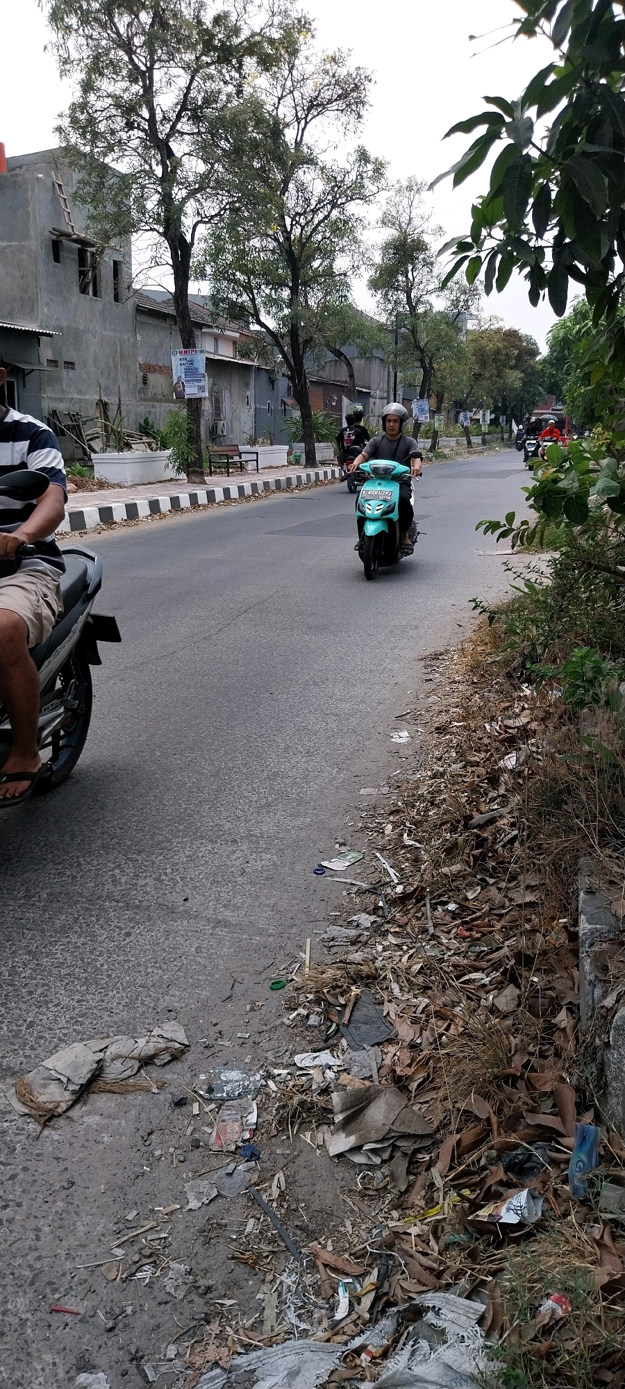 The image depicts a street scene in Bojong Nangka, Indonesia. The foreground is dominated by a paved road with a littered shoulder on the right, featuring dry leaves, discarded plastic, and other debris. On the left side of the road, a person wearing a striped shirt and shorts is seated on a motorcycle, partially visible. Further down the road, a person wearing a helmet and a grey t-shirt is riding a turquoise scooter. Behind them, other motorcycles are visible in the distance. The background shows a row of houses and trees lining the street. The sky is overcast.