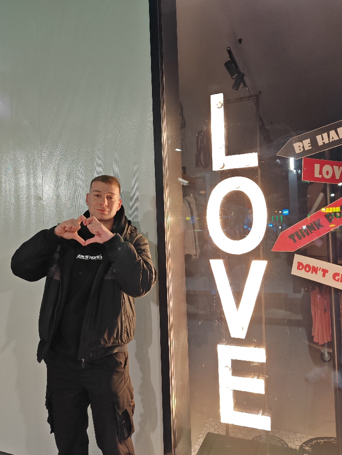 A young adult male, with a light skin tone and a short buzz-cut, is captured standing outdoors in the city of Shumen, Bulgaria. He is positioned slightly left of center, facing directly towards the camera, and holds both hands in front of his chest, forming a heart shape with his thumbs and index fingers. The individual is dressed in a black hooded sweatshirt with the word "EASTGATE" visible on the chest, a black puffer jacket worn over the sweatshirt, and black cargo pants. To his right, a large retail storefront window displays prominent vertical illuminated white letters spelling "LOVE." Within the store window, several arrow-shaped signs with white text on dark red/brown backgrounds are visible, reading "BE HAPPY," "LOVE," "THINK POSITIVE," and "DON'T GIVE UP." Reflections of distant urban lighting indicate an evening or nighttime setting. Clothing racks with light-colored garments are faintly discernible inside the store through the window. A plain, light grey wall serves as the background to the man's left.