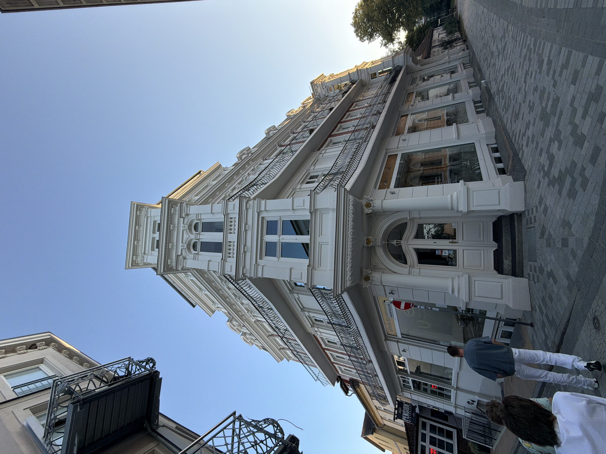 The image depicts a street corner in Rostock, Germany, featuring a grand, white, multi-story building with ornate architectural details, including balconies with wrought-iron railings and decorative cornices. The building's facade is angled, creating a dramatic perspective. On the ground floor, large windows suggest commercial establishments. Two people are visible walking away from the camera on a paved sidewalk. A sign for "Pension" is partially visible on an adjacent building. The sky is clear and blue, indicating daylight. The overall scene conveys a sense of European urban architecture.