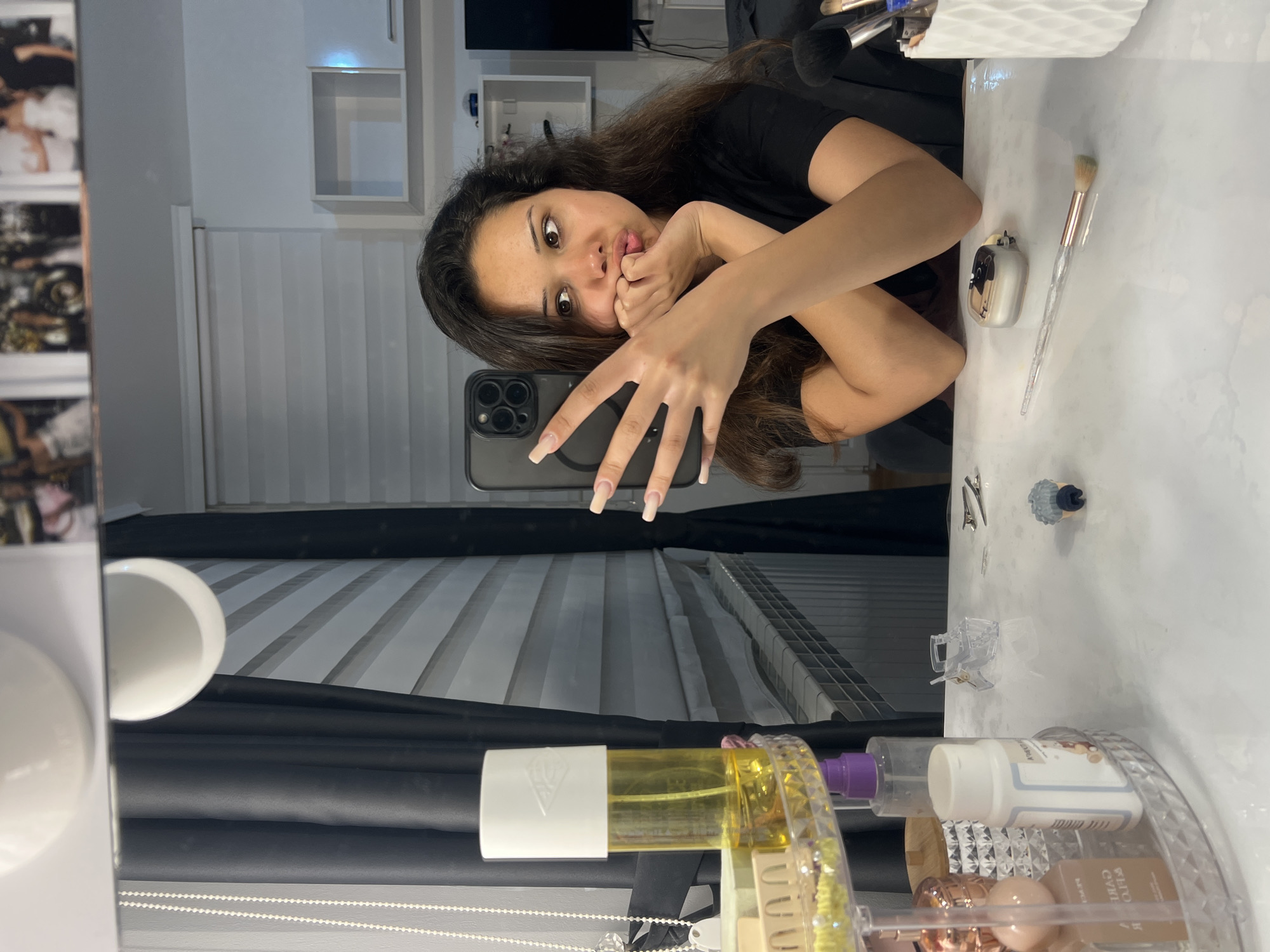 In a brightly lit room, a young woman with long, dark hair is reflected in a mirror. She is seated at a vanity table, her arms crossed and her hands touching her chin as she looks off to the side with pursed lips. She is wearing a black t-shirt and has long, light-colored artificial nails. On the vanity, various makeup products and accessories are arranged, including a tall yellow spray bottle, a white pill bottle, a clear rotating organizer, and several makeup brushes in a white holder. In the background, through the mirror's reflection, blinds cover a window, and shelves are visible on the wall. The scene appears to be a personal dressing area, likely within a residence in Zlatograd, Bulgaria, given the context.