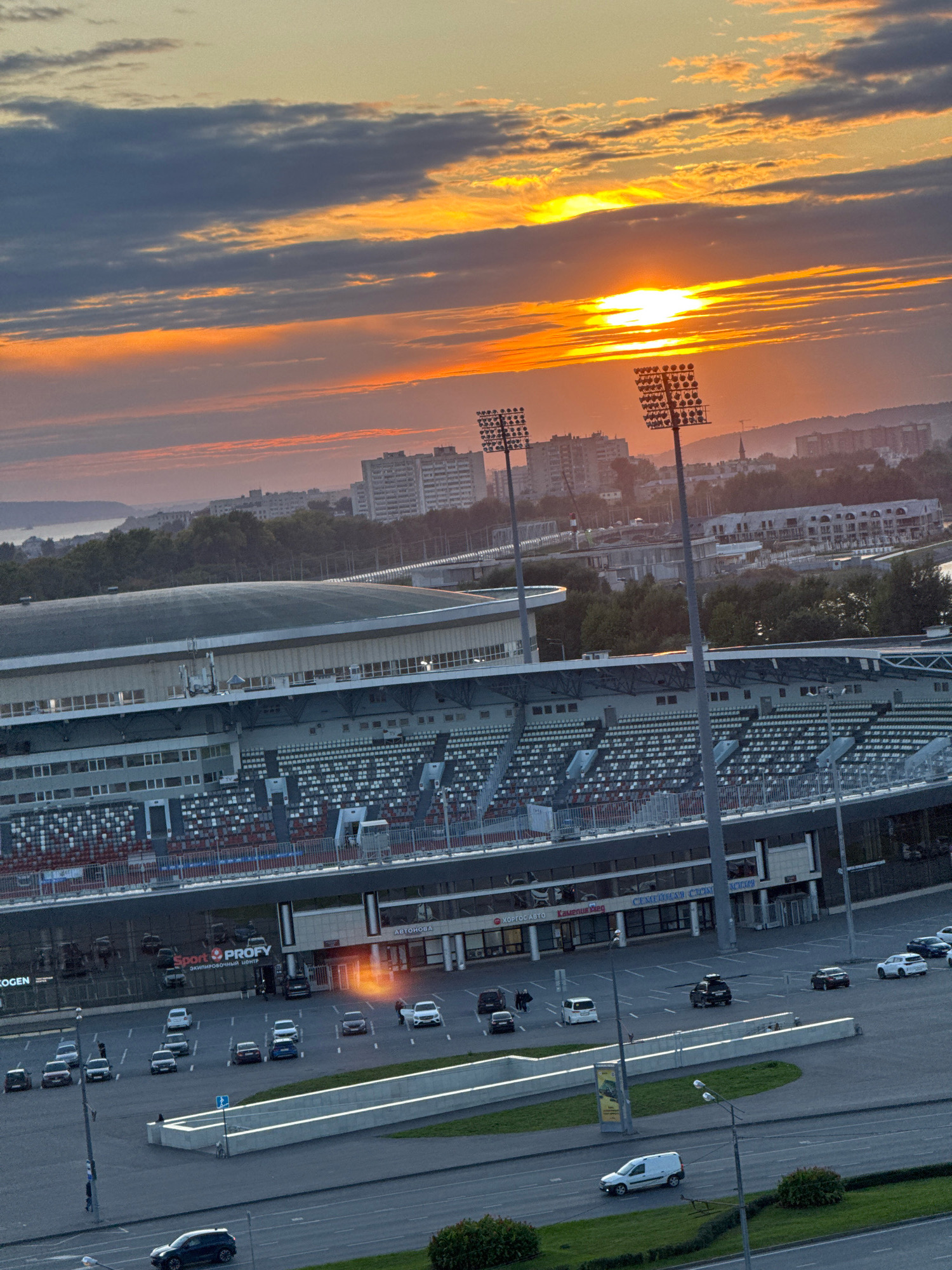 The image captures a panoramic view of a stadium in Kazan, Russia, during a sunset. The sky is ablaze with orange and yellow hues, with scattered clouds creating a dramatic backdrop. The stadium, with its tiered seating and prominent floodlights, dominates the midground. In the foreground, a parking lot is visible with several cars. The overall scene is characterized by the interplay of natural light and urban infrastructure.