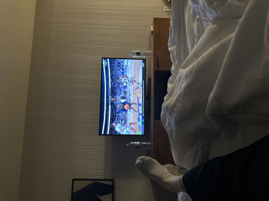 A person lies in bed watching a basketball game on television. The bed is covered in white sheets and a comforter. The person is wearing socks and dark pants. Two water bottles sit on the dresser to the side of the television. The wall has a lined wallpaper and a framed piece of art is visible.