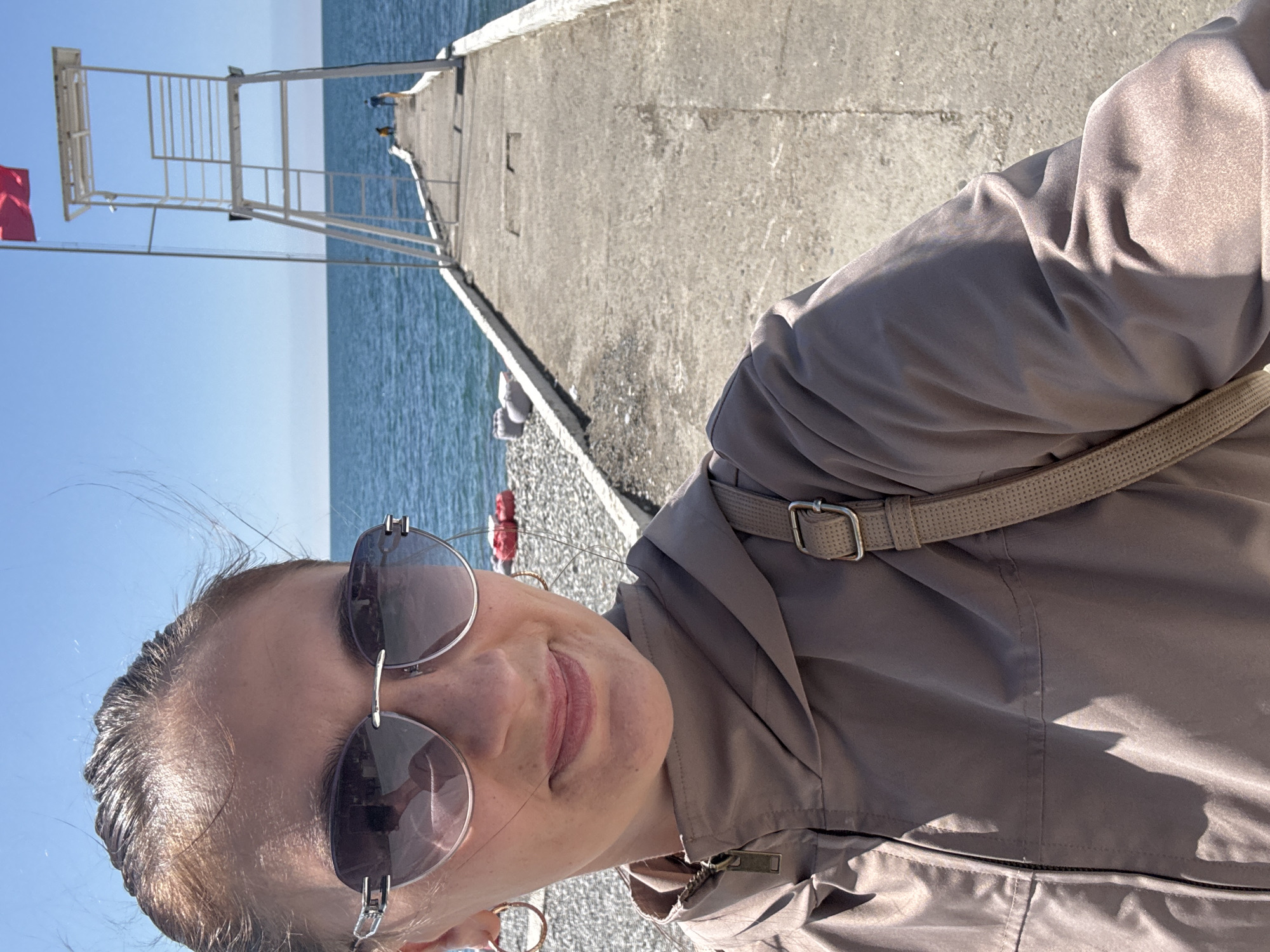 A woman wearing sunglasses and a gray jacket takes a selfie on a pier in Sochi, Russia. Behind her is the Black Sea, a metal lifeguard tower, and a flag. The pier is made of concrete.