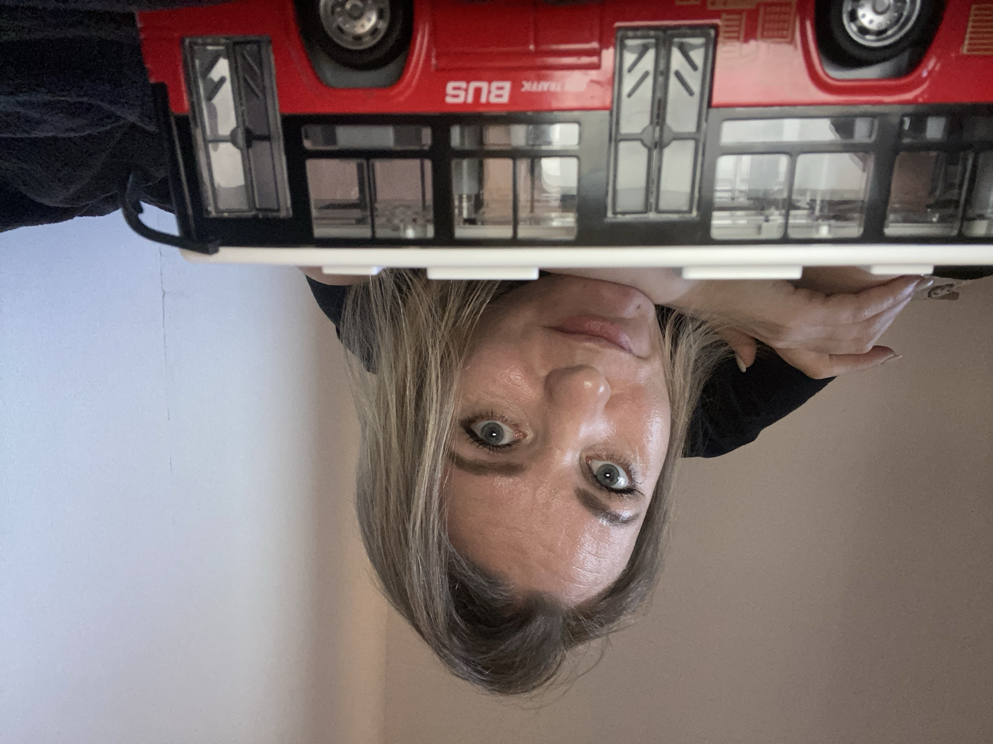The image features a woman looking directly upwards towards the viewer, with her head and upper torso visible. She has light-colored, possibly blonde or light brown, hair and wide, striking blue-green eyes with dark eyeliner, conveying an expression of surprise or curiosity. Her face appears slightly shiny under the indoor lighting. She is wearing a dark-colored top, and her hands are partially visible near her head on the right side of the frame.
Positioned above the woman's head is a red toy model of a double-decker bus. The bus is seen from its side, with black window frames and visible wheels. White text printed on its side clearly reads "BUS," and beneath it, partially visible and reversed, the word "TRAFFIC" can be discerned. The bus appears to be resting on a flat, light-colored surface directly above the woman. A dark, textured fabric, possibly clothing or a blanket, is partially visible on the far left of the image, near the bus.
The background consists of plain walls, which are a light off-white or grey on the left and a warmer beige or tan on the right, indicating an indoor setting. The image is taken from a low angle, suggesting the woman is looking up at the object or camera. While the location context is Saint Petersburg, Russia, there are no specific visual cues within the image itself to confirm this.