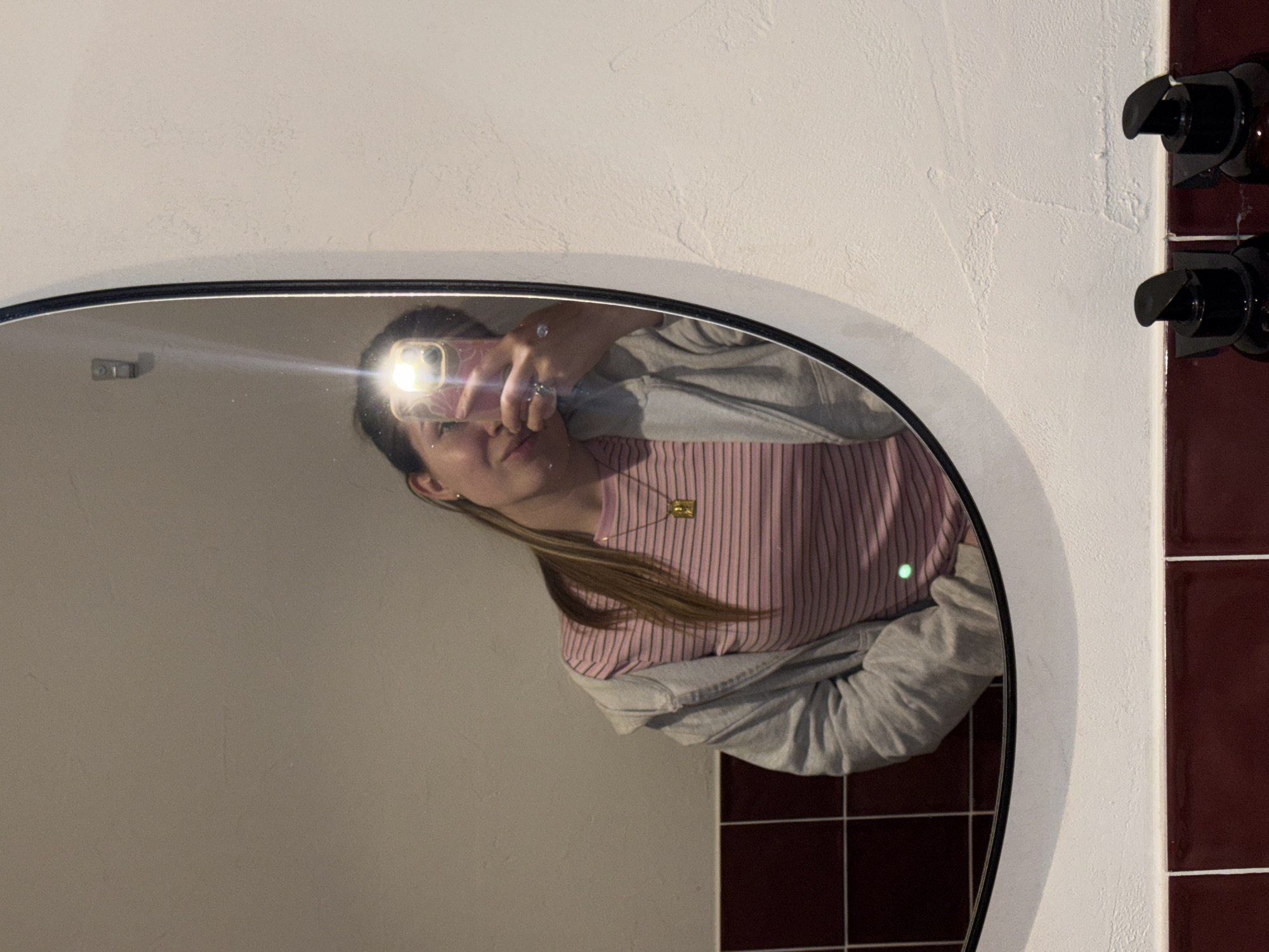 A person is captured in a mirror selfie, standing in what appears to be a bathroom. The subject, a young individual with long brown hair, is wearing a pink and white striped t-shirt layered under a grey hoodie or sweatshirt. They are also wearing a gold-colored rectangular pendant necklace and a ring on one finger. Their face is partially obscured by the smartphone they are holding, which is emitting a bright flash that creates a prominent glare in the mirror.
The mirror itself is oval or irregularly shaped with a thin black frame. Reflected within the mirror are sections of a white textured wall and dark reddish-brown square tiles. To the immediate right of the mirror, two black pump dispensers are mounted vertically on the white wall, positioned above a strip of the dark reddish-brown tiles. A small green light dot is visible on the person's chest area in the reflection. The image is oriented with the top of the mirror to the right, implying a 90-degree counter-clockwise rotation from a typical upright view. The overall scene suggests an indoor setting, likely a private space such as a bathroom, located in Belgrade, Serbia. No visible text is present in the image.