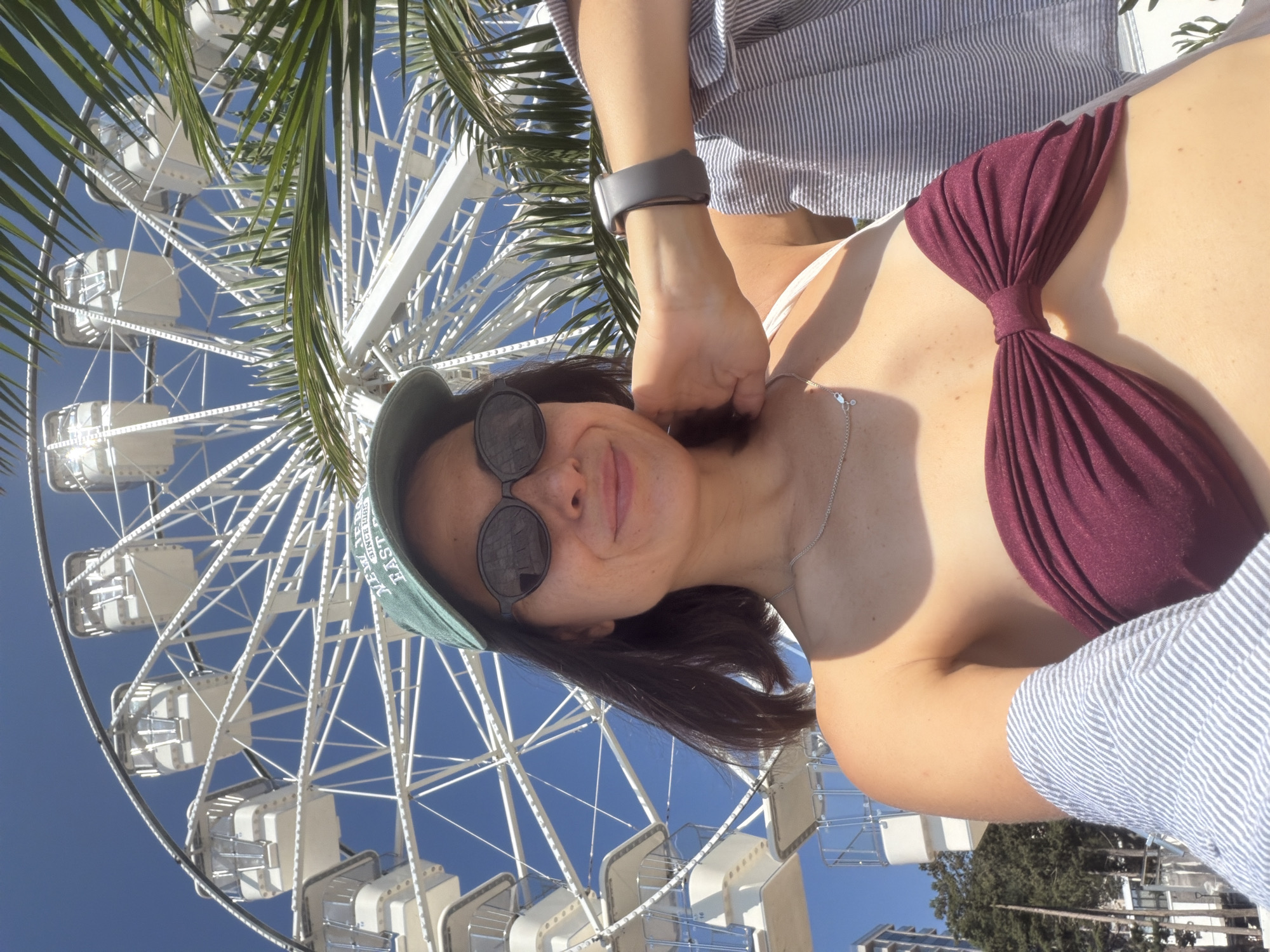 A person is taking a selfie in front of a Ferris wheel. The person is wearing sunglasses, a green baseball cap, and a burgundy bikini top. They are smiling at the camera. The Ferris wheel is white and appears to be in operation, with its gondolas visible against a clear blue sky. Palm fronds are visible in the foreground, partially obscuring the view of the Ferris wheel. The image appears to have been taken during the daytime, likely in a sunny climate. The overall mood of the image is cheerful and vacation-like. There is text visible on the green baseball cap which appears to read "EAST SINCE".