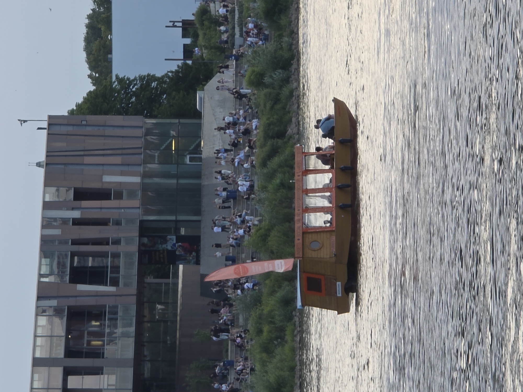 The image captures a wide shot of a riverfront scene in Warsaw, Poland. In the foreground, a small wooden boat with a red sail is traversing the rippling water. The boat appears to be a passenger vessel, with several people visible inside, some seated and others standing. On the bank of the river, a large, modern building with extensive glass facades is prominent. This building overlooks a tiered, amphitheater-like structure where a crowd of people are gathered, some sitting on the steps and others standing. Lush green trees and vegetation line the riverbank between the water and the amphitheater. The sky above is overcast, suggesting it might be late afternoon or evening. The overall atmosphere suggests a public gathering place and recreational area in the city.