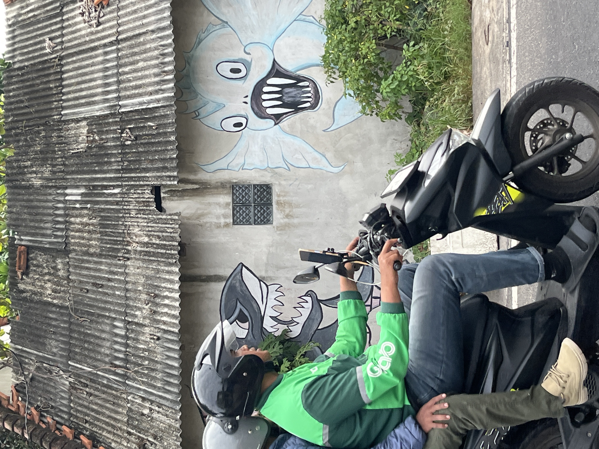 A motorcyclist wearing a green jacket and a black helmet rides a scooter on a street in Kuta Selatan, Indonesia. A passenger wearing a grey helmet is seated behind the driver. The scooter is parked in front of a wall adorned with murals of a shark and a light blue, cartoonish fish with its mouth open. The wall is under a corrugated metal roof. Green foliage is visible behind the wall. The street appears to be paved with asphalt.