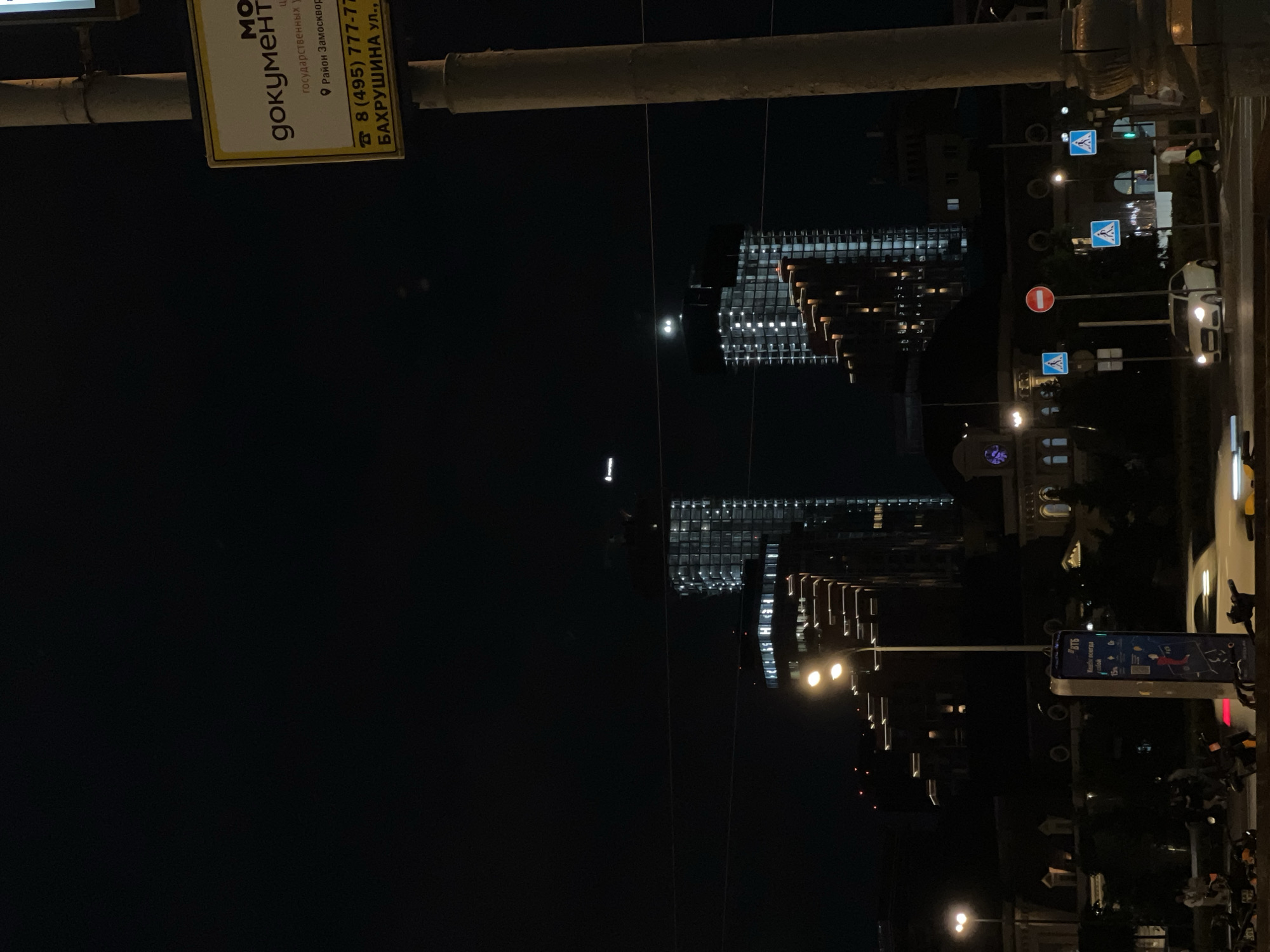 A nighttime street scene in Moscow, Russia. Two modern buildings, illuminated with rows of lights, stand tall against the dark sky. A street sign displays “МО документ” indicating government services, with the address “БАХРУШИНА ул.” and phone number “8 (495) 777-77”. Streetlights line the road, and pedestrian crossing signs are visible. A VTB bike rental station is present, and several parked motorcycles are on the sidewalk. The moon is faintly visible in the sky.
