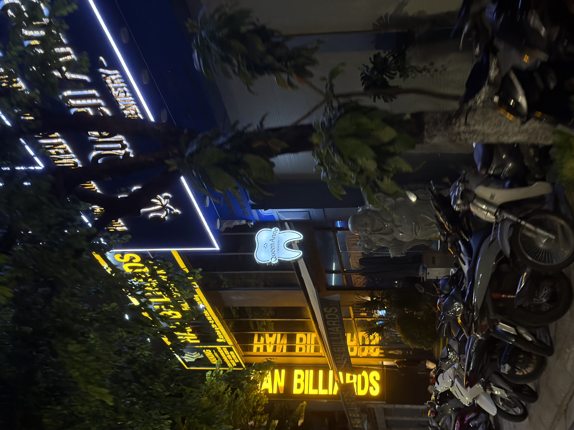 The image shows a nighttime street scene in Da Nang, Vietnam, featuring illuminated signs for "QUEEN ANNE DENTISTRY" and "HAN BILLIARDS". The dentistry sign is a bright white neon outline of a tooth with the text "Queen Anne" and "DENTISTRY" inside. Above it, a large blue sign with illuminated white and yellow text is visible. The "HAN BILLIARDS" sign is vertically oriented, with bright yellow neon lettering. Below the signs, a row of parked motorcycles is visible. A statue of a Buddha is also present near the entrance of the building. Trees with lush foliage obscure parts of the scene. The street appears to be well-lit by the neon signs and ambient city lights.
