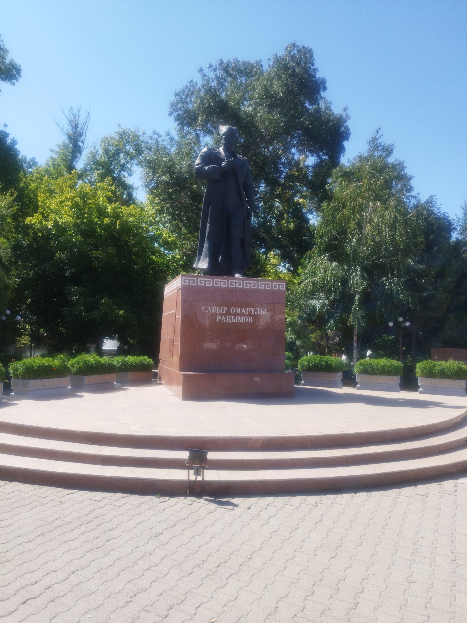 The image shows a statue of a man in formal attire, possibly a historical figure or a politician, standing on a red granite pedestal. The inscription on the pedestal reads "САБЫР ОМАРУЛЫ РАКЫМОВ". The statue is located in an outdoor public space with a paved walkway and several tiers of steps leading up to a raised platform. There are decorative planters with green foliage around the platform, and trees form a backdrop to the scene. The sky is clear and blue, indicating a sunny day. Based on the inscription and the context of similar statues, this is likely the statue of Sabir Omirulievich Rakhimov, a Soviet military leader, located in Shymkent, Kazakhstan.