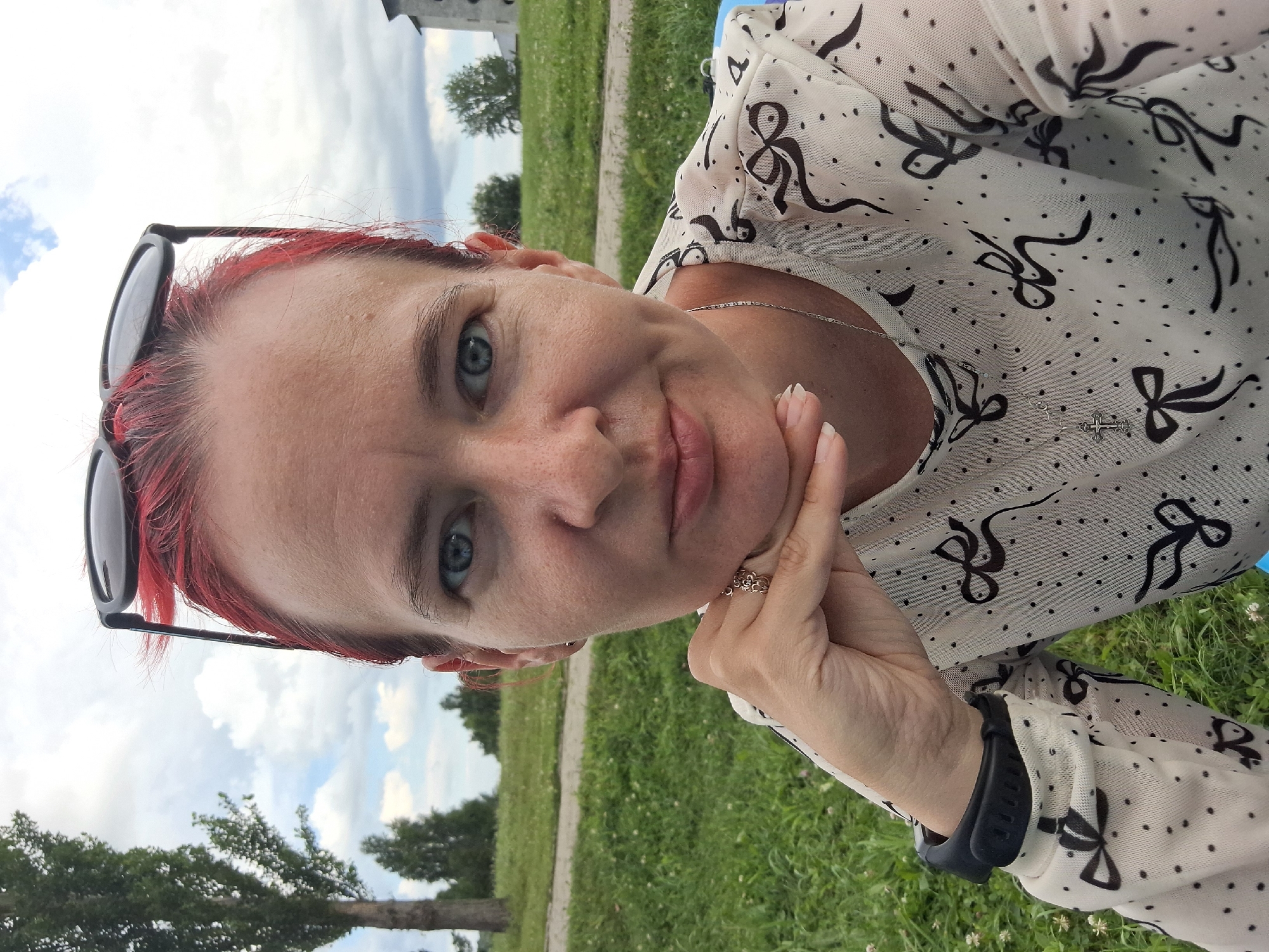 A woman with bright red hair pulled back and sunglasses perched on her head is captured in a close-up, frontal selfie. Her blue eyes are looking directly at the camera, and her chin rests on her left hand, which is adorned with a floral-patterned ring. She is wearing a white blouse with a repeating black bow and dot pattern, and a silver cross necklace. A black watch is visible on her left wrist. The background suggests an outdoor setting with a grassy area, a path, and trees, possibly in a park or rural area. The sky is partly cloudy. There is no visual information in the image to indicate the location is the city of Atamanovka, Russia.