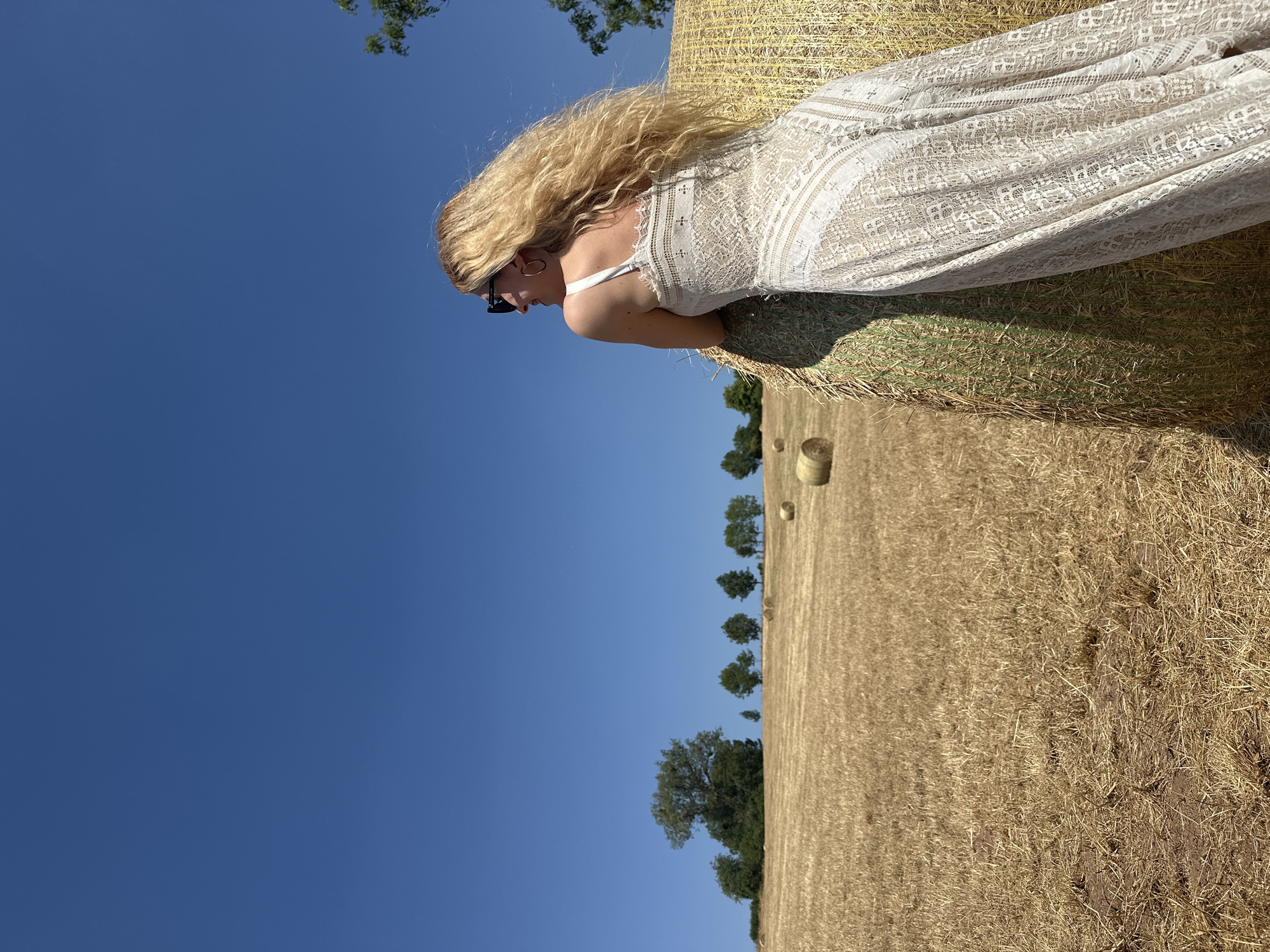 In Tandel, Luxembourg, a blonde woman wearing a white lace dress and sunglasses leans against a large, round hay bale in a sunlit, harvested field. The sky above is a clear, vibrant blue, with a few distant trees visible on a gentle slope in the background. Several other hay bales are scattered across the dry, golden field, suggesting it's late summer or early autumn. The woman is facing away from the camera, looking out towards the horizon, her long, wavy blonde hair blowing in the breeze. The scene evokes a sense of rural tranquility and a bright, warm day.