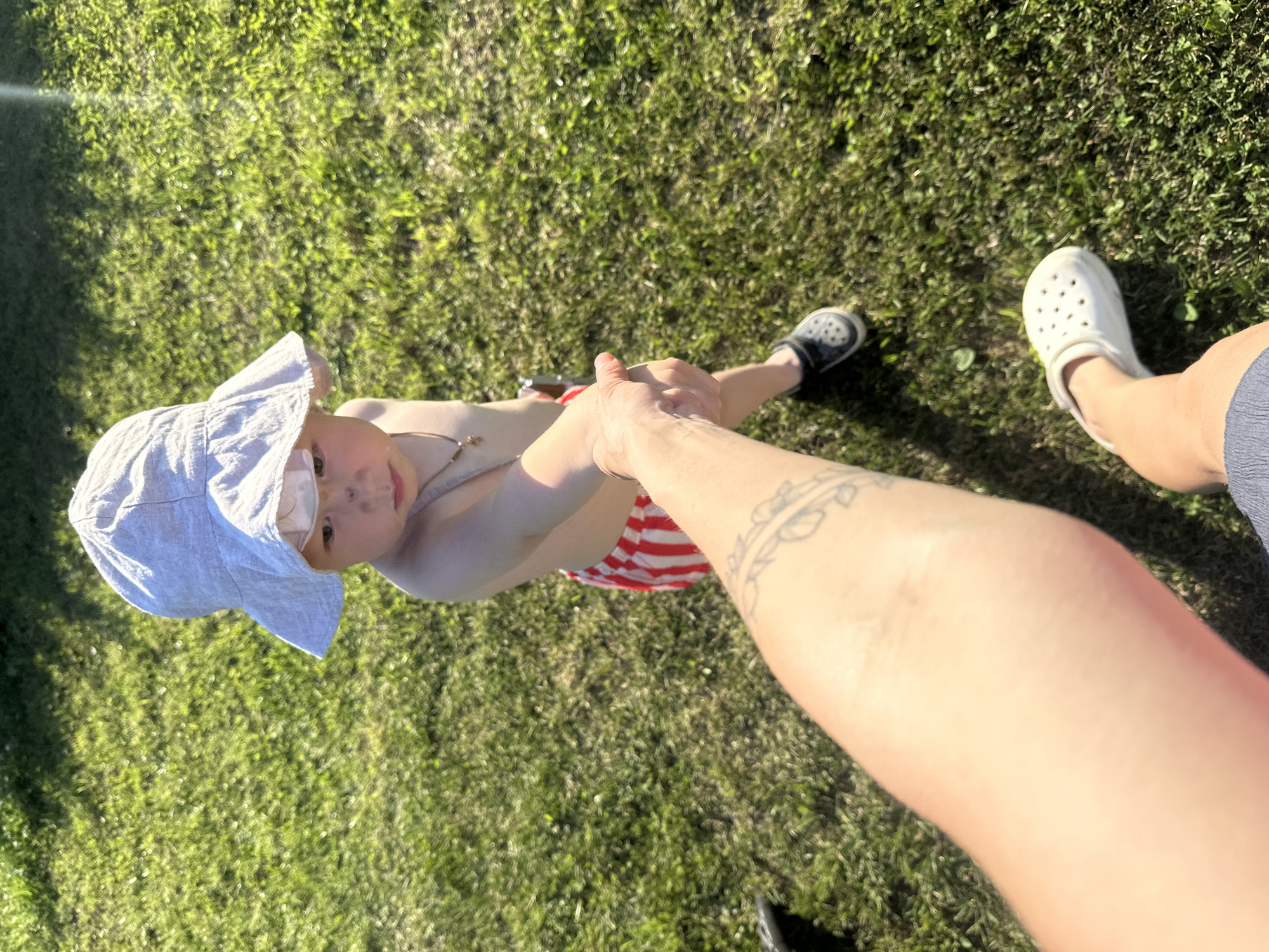 The media file shows a high-angle shot of two people on a grassy lawn. One person, a child wearing a light gray sun hat and red and white striped shorts, is held by the hand by the other person, whose arm and leg are visible in the foreground. The child is looking up towards the camera with a neutral expression. The other person is wearing a white Croc shoe and has a tattoo on their arm. The scene appears to be outdoors on a sunny day, with the sunlight casting shadows on the grass. The overall impression is of a casual, outdoor activity, possibly a child playing with an adult. The location context provided is Galyevo, Russia. There is no visible text in the image.