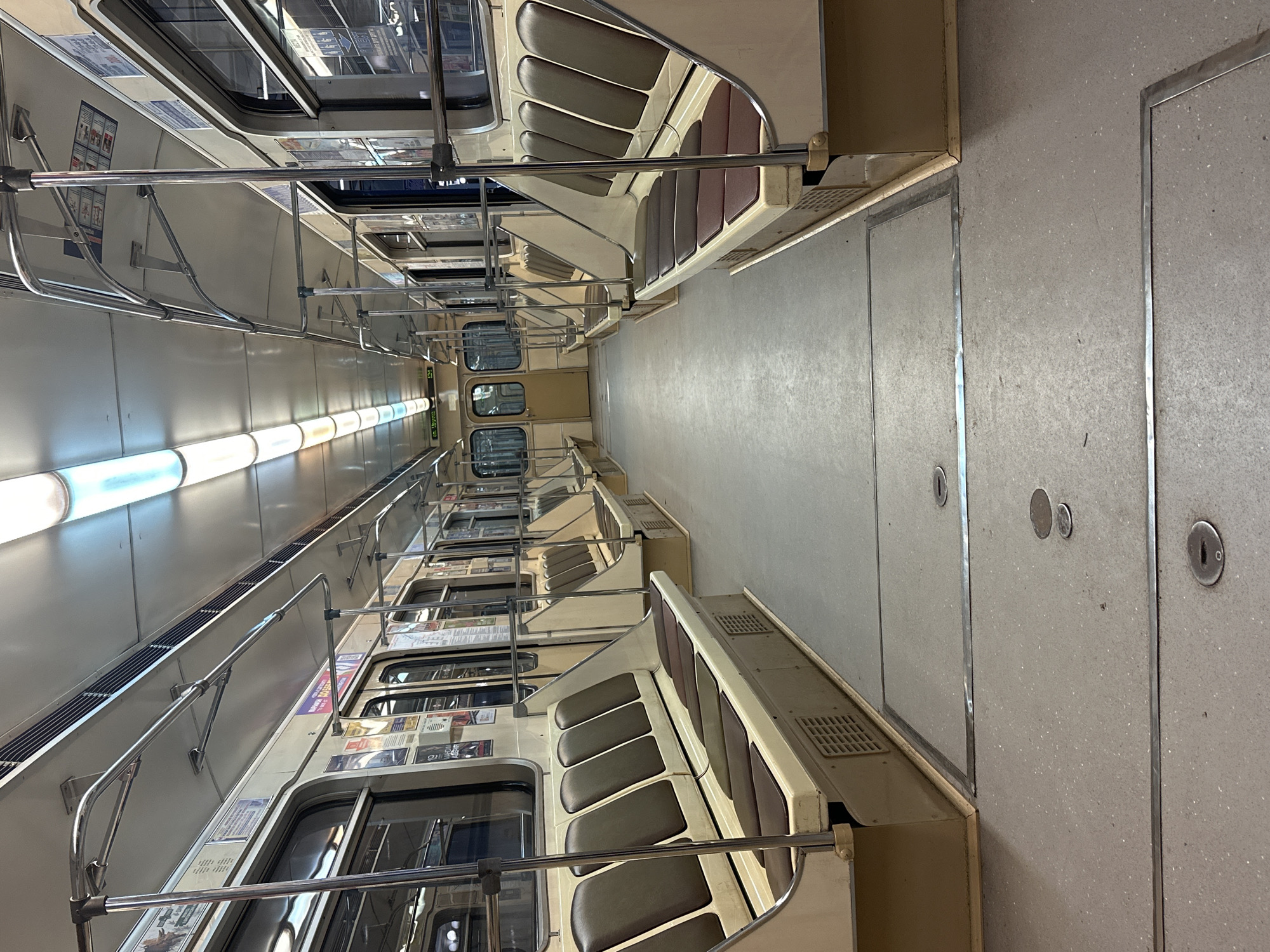 The image shows the interior of an empty subway car, likely in Minsk, Belarus, given the Cyrillic text visible on posters. The car is clean and well-lit by fluorescent lights. The seats are upholstered in a brown material and are arranged in a forward-facing configuration along the sides of the car. Handrails made of metal are mounted on the ceiling and along the sides of the car. The floor is a light gray, speckled material with metal strips marking sections. The overall impression is one of a functional and utilitarian public transportation vehicle.