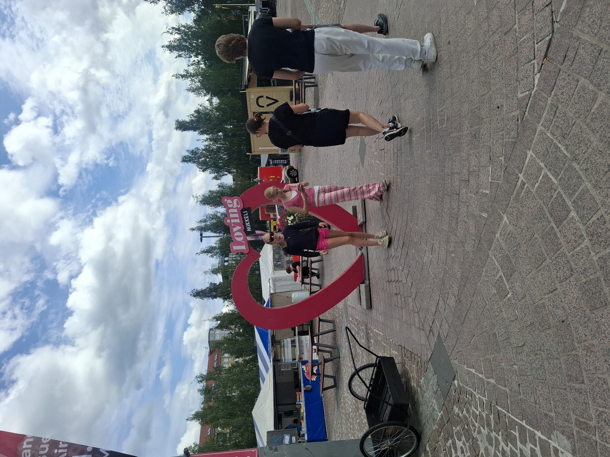 The image depicts an outdoor public square in Mikkeli, Finland, characterized by cobblestone pavement and a cloudy sky. Dominating the center of the scene is a large, bright pink heart sculpture with the word "Loving" visible on its surface, along with "MIKKELI" below it.
Several individuals are present in the square. To the left of the heart, a young girl with blonde hair pulled back wears pink shorts and a sleeveless top, looking down at her phone. Next to her, a woman with dark hair and glasses wears a black dress. Further to the right, a person with short, light brown hair, wearing a black t-shirt and white pants, walks away from the camera. In the background, a food stall with a blue and white striped awning is set up.
To the left of the heart sculpture, a black wheeled cart with a wooden bed is positioned on the cobblestones. The environment appears to be a marketplace or a public gathering space, suggested by the presence of stalls and picnic tables in the background. The overall atmosphere is casual, with people engaged in various activities in a public setting.
